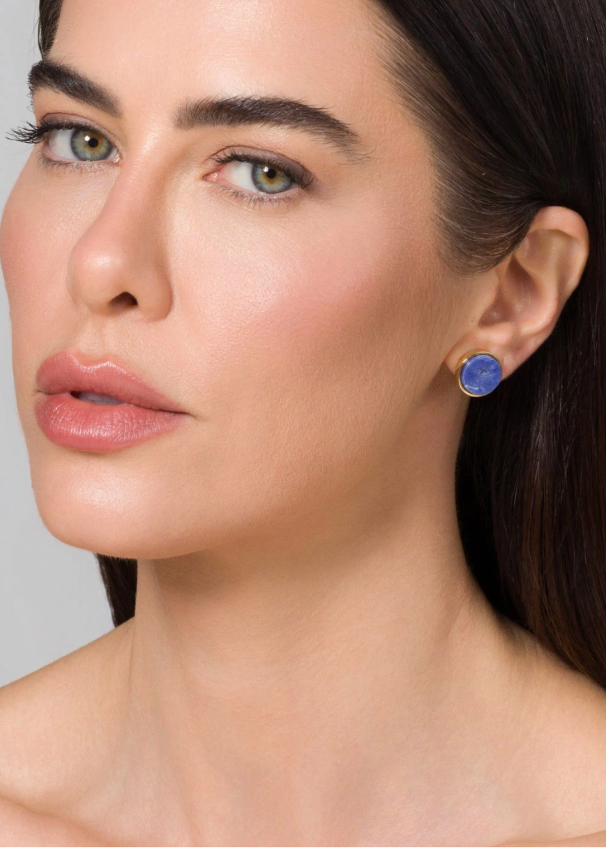 Close-up of a woman wearing a small round blue stud earring with a gold-toned bezel setting against a light gray background.