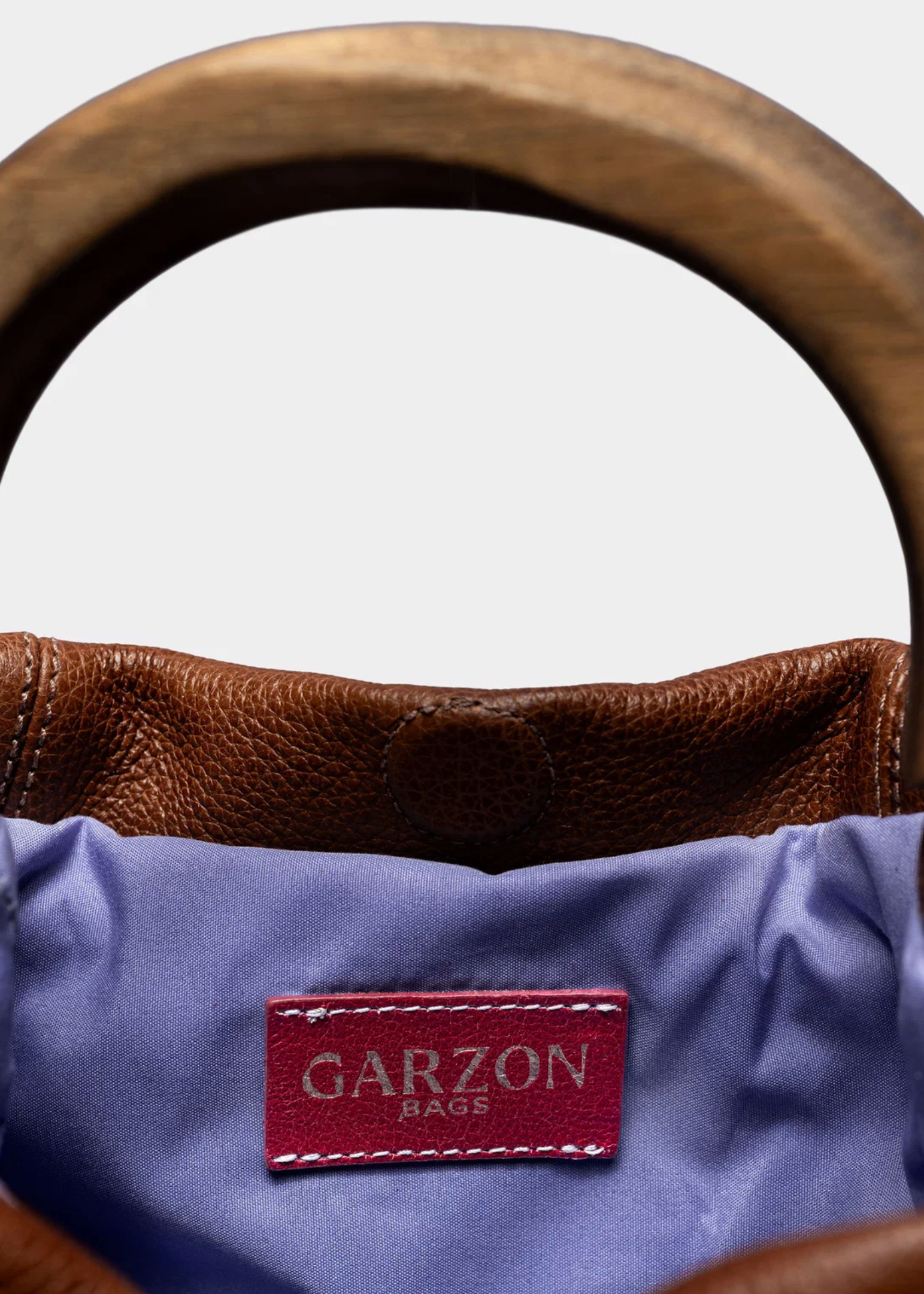 Close-up of a brown leather handbag with a purple interior lining and a red GARZON BAGS brand patch, framed by a wooden handle against a light gray background.