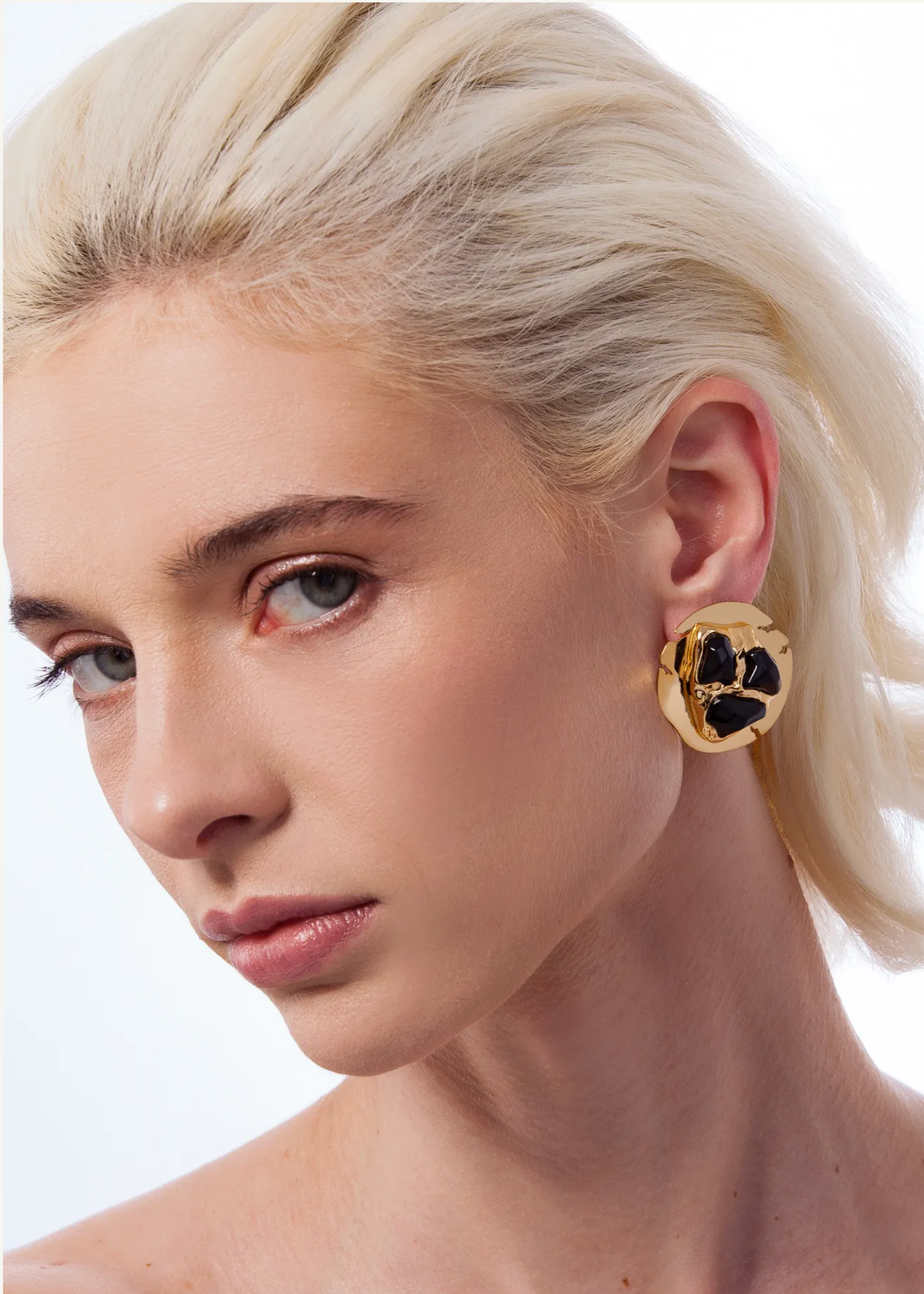 A close-up portrait of a blonde woman wearing a gold-toned statement earring with black floral detailing, set against a clean white background.