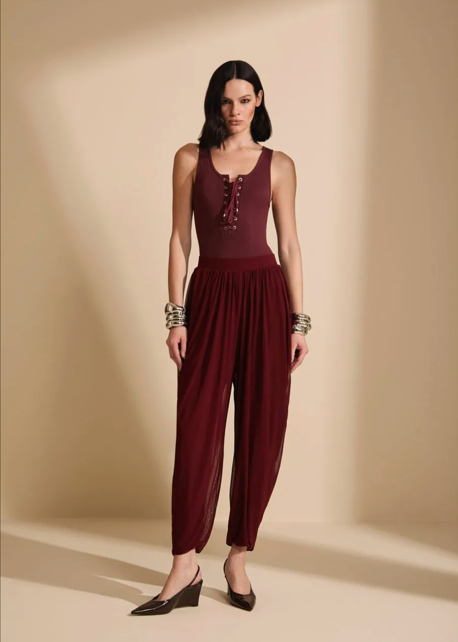A woman modeling a sleeveless burgundy jumpsuit with a lace-up front and loose harem-style pants, standing against a warm beige studio backdrop.