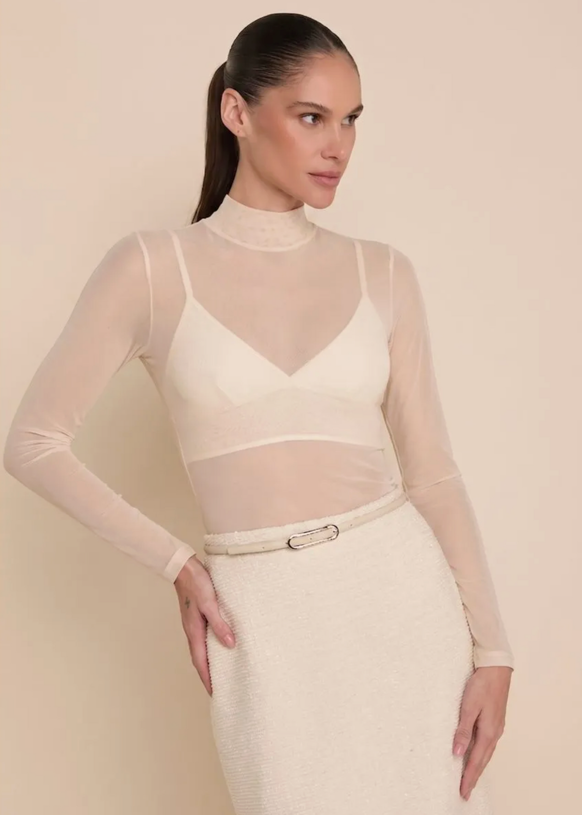 A woman modeling a cream-colored sheer long-sleeve mock neck top with a bralette overlay and a matching textured cream skirt, set against a solid beige studio background.
