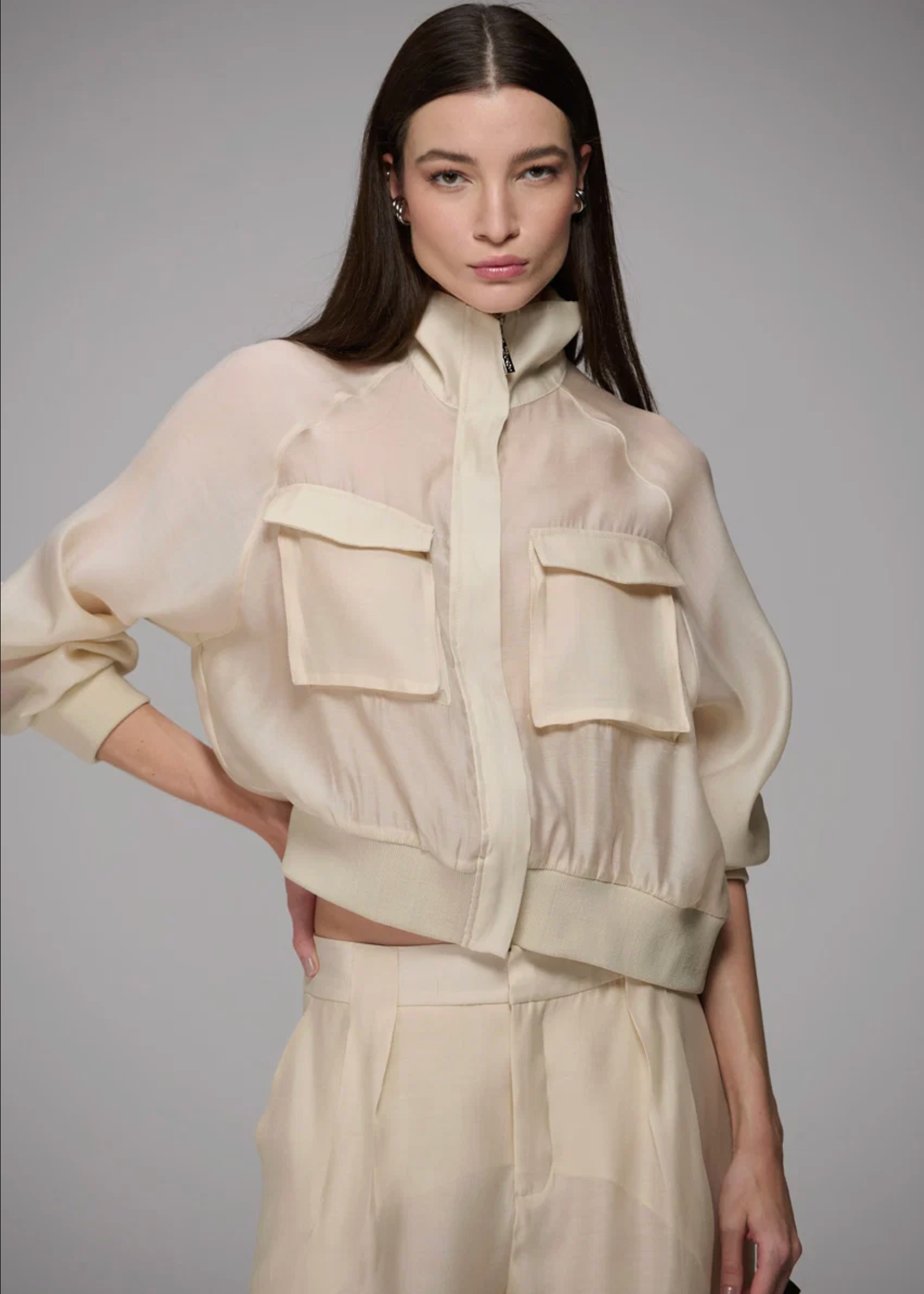 A model wearing a cream-colored semi-sheer cropped jacket with large front flap pockets and matching high-waisted trousers, set against a solid gray studio background.