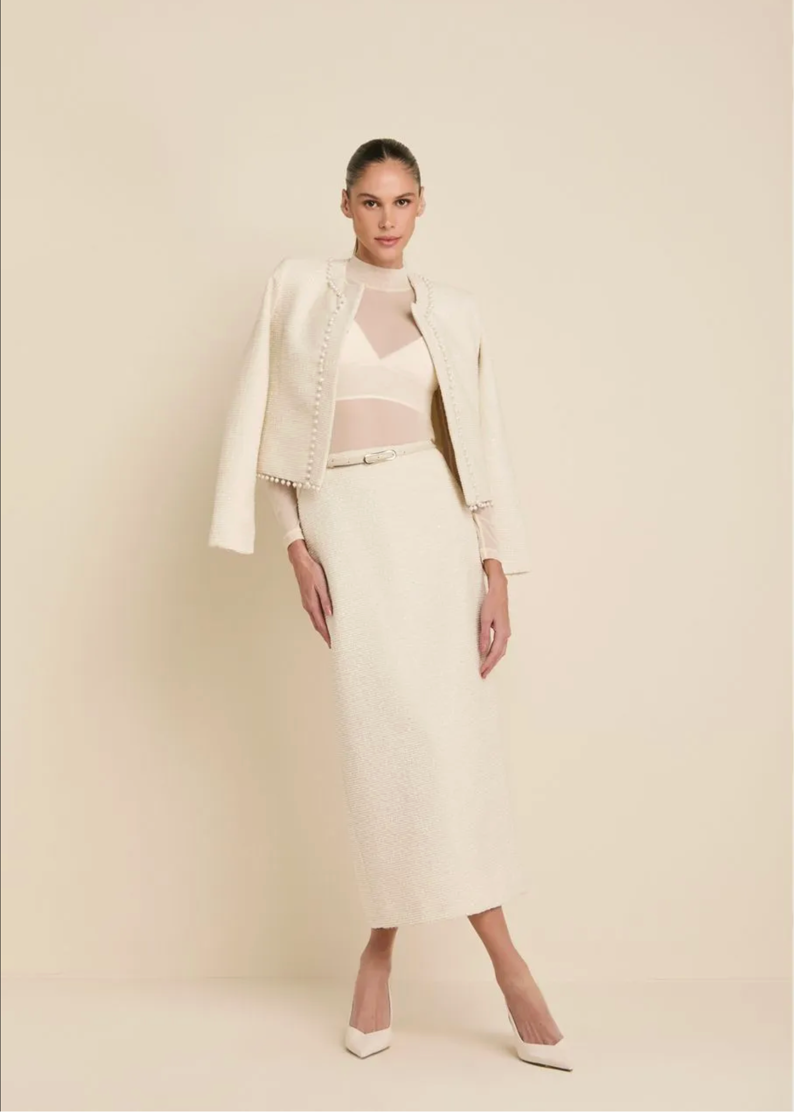 A model stands against a warm beige studio background wearing a coordinated cream outfit consisting of a textured midi skirt, a matching cropped jacket, and a sheer mesh top with white pointed heels.