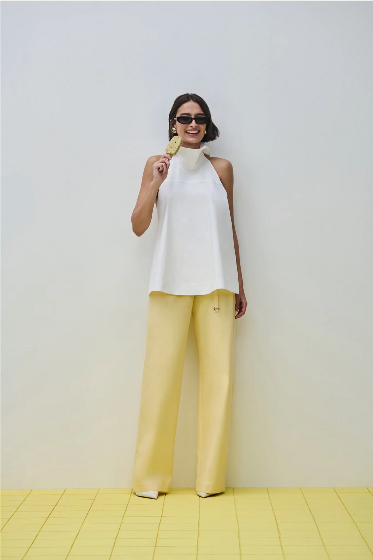 A woman modeling a sleeveless white mock-neck top and pale yellow wide-leg trousers, standing against a light gray wall on a yellow tiled floor.