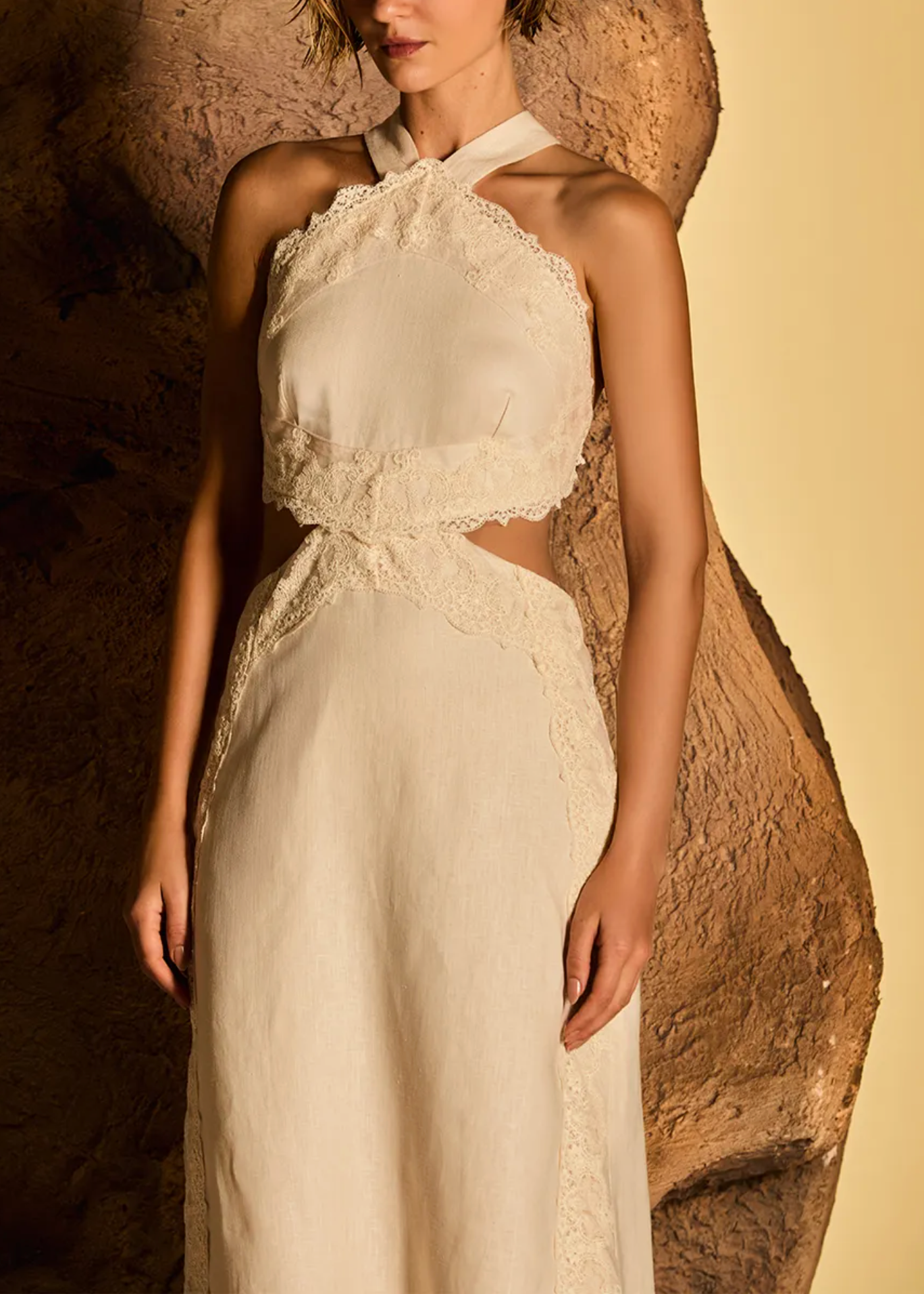 A model wears a cream-colored halter-neck dress with intricate lace trim and side cutouts, posed against a textured brown rock and a warm beige background.