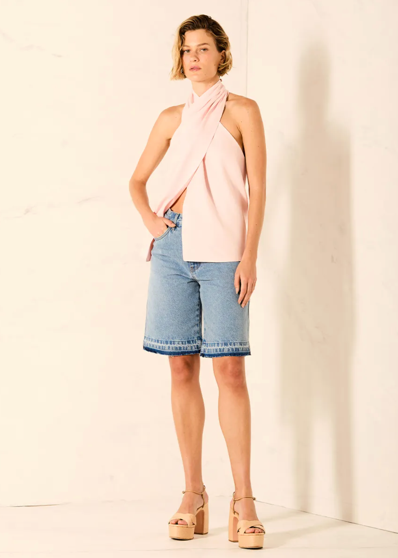 A woman models a light pink asymmetrical draped halter top paired with light-wash denim shorts and beige platform sandals, standing against a white marble-textured wall.
