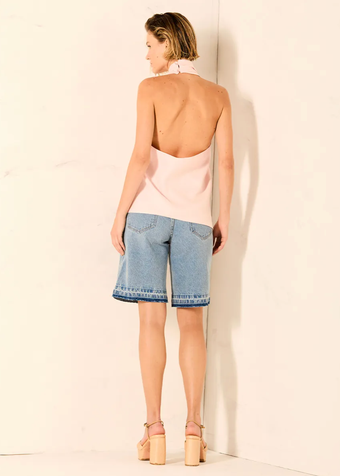 A woman stands with her back to the camera wearing a pale pink halter-style backless top, light blue denim shorts, and beige platform sandals against a plain off-white wall.