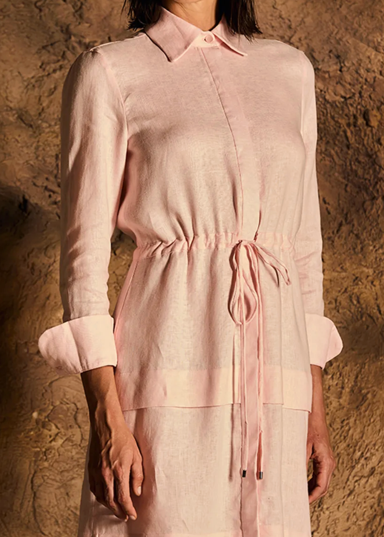 A model wearing a light pink linen shirt dress with a collar, button-down front, three-quarter sleeves with rolled cuffs, and a drawstring waist, posed against a textured brown rock background.