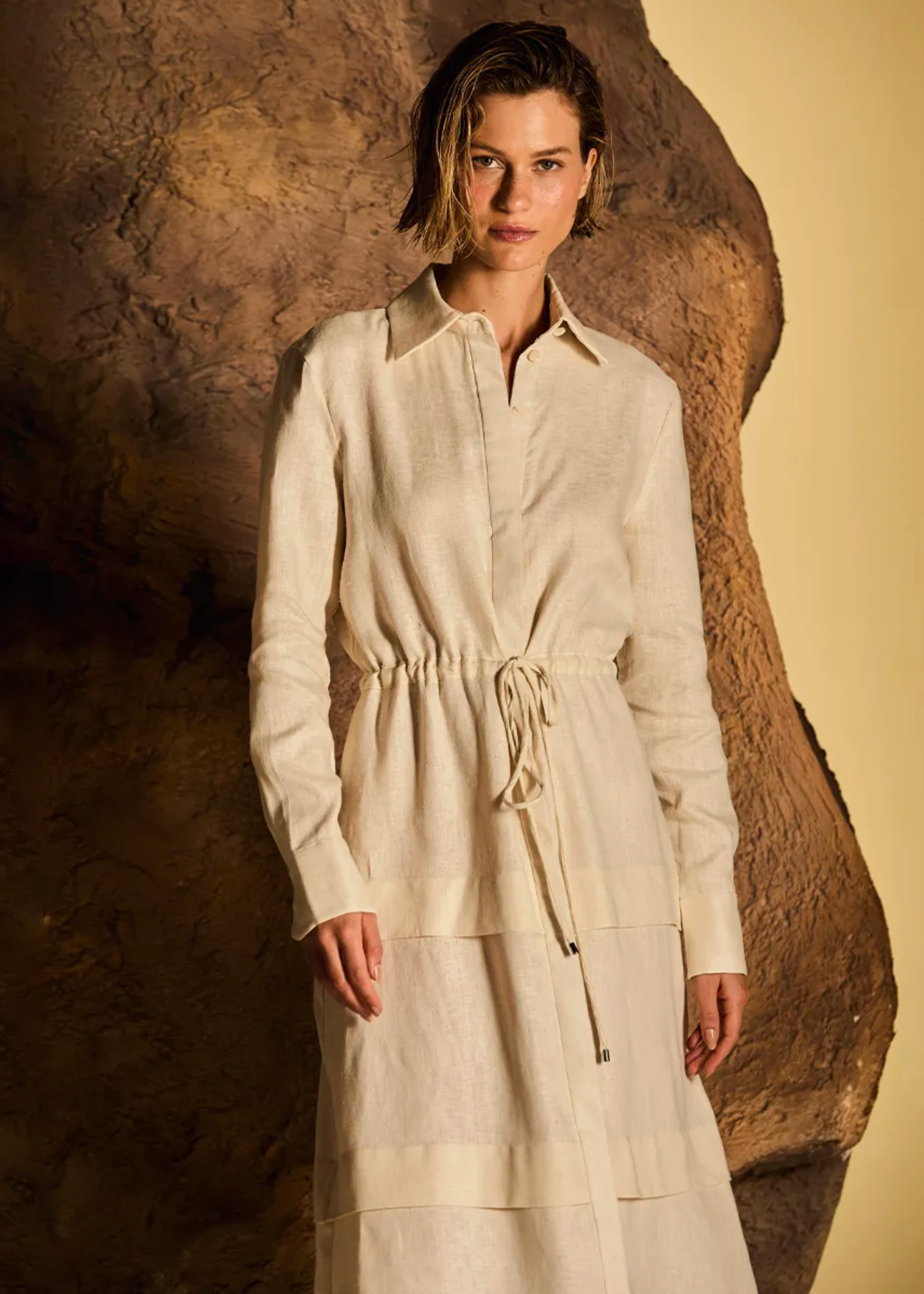 A woman modeling a long-sleeve cream-colored linen shirt dress with a drawstring waist, standing against a textured brown rock and a warm beige background.