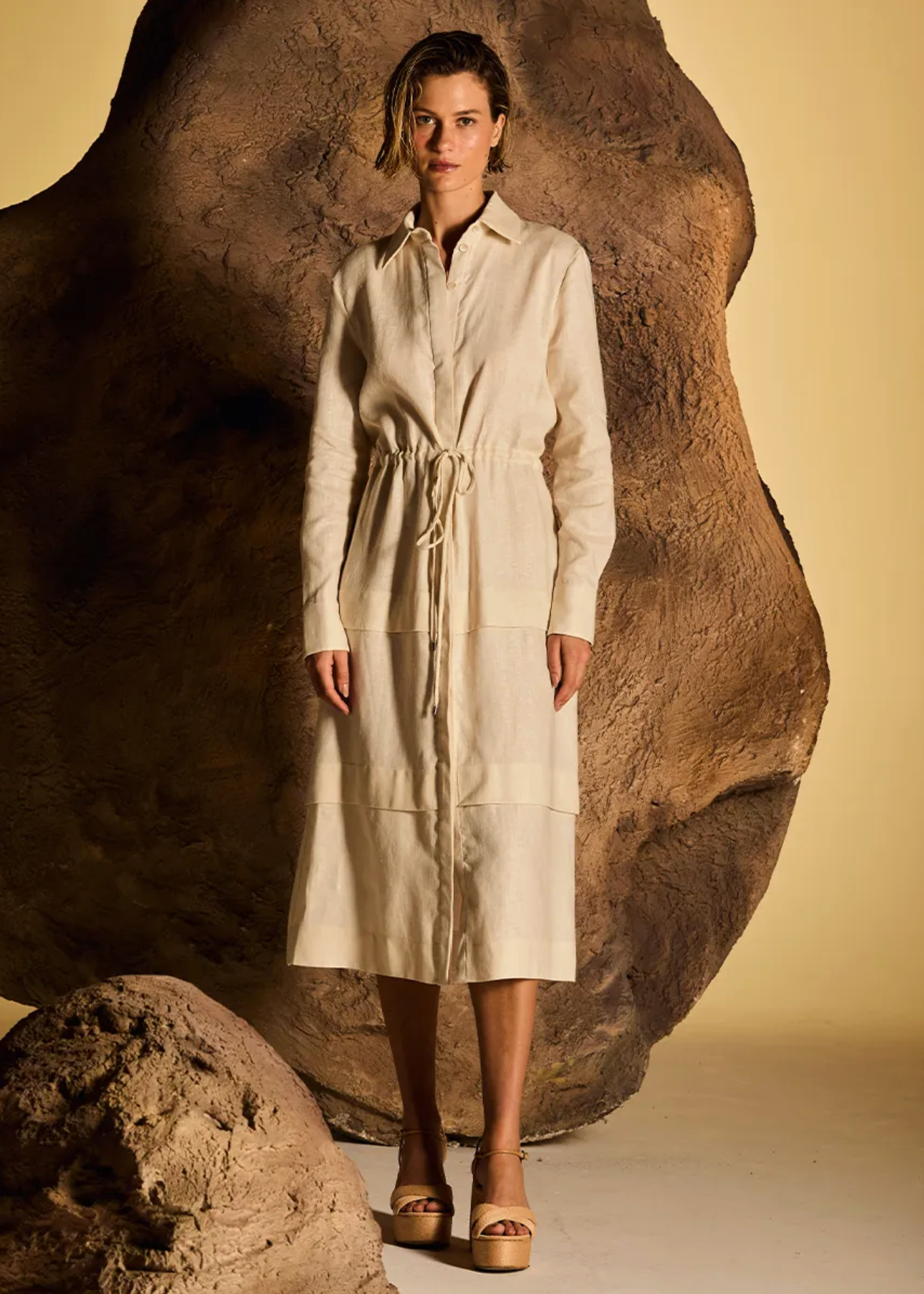 A woman stands in a studio wearing a long cream-colored shirt dress with a tie waist, posed in front of a large sculptural rock against a warm beige background.