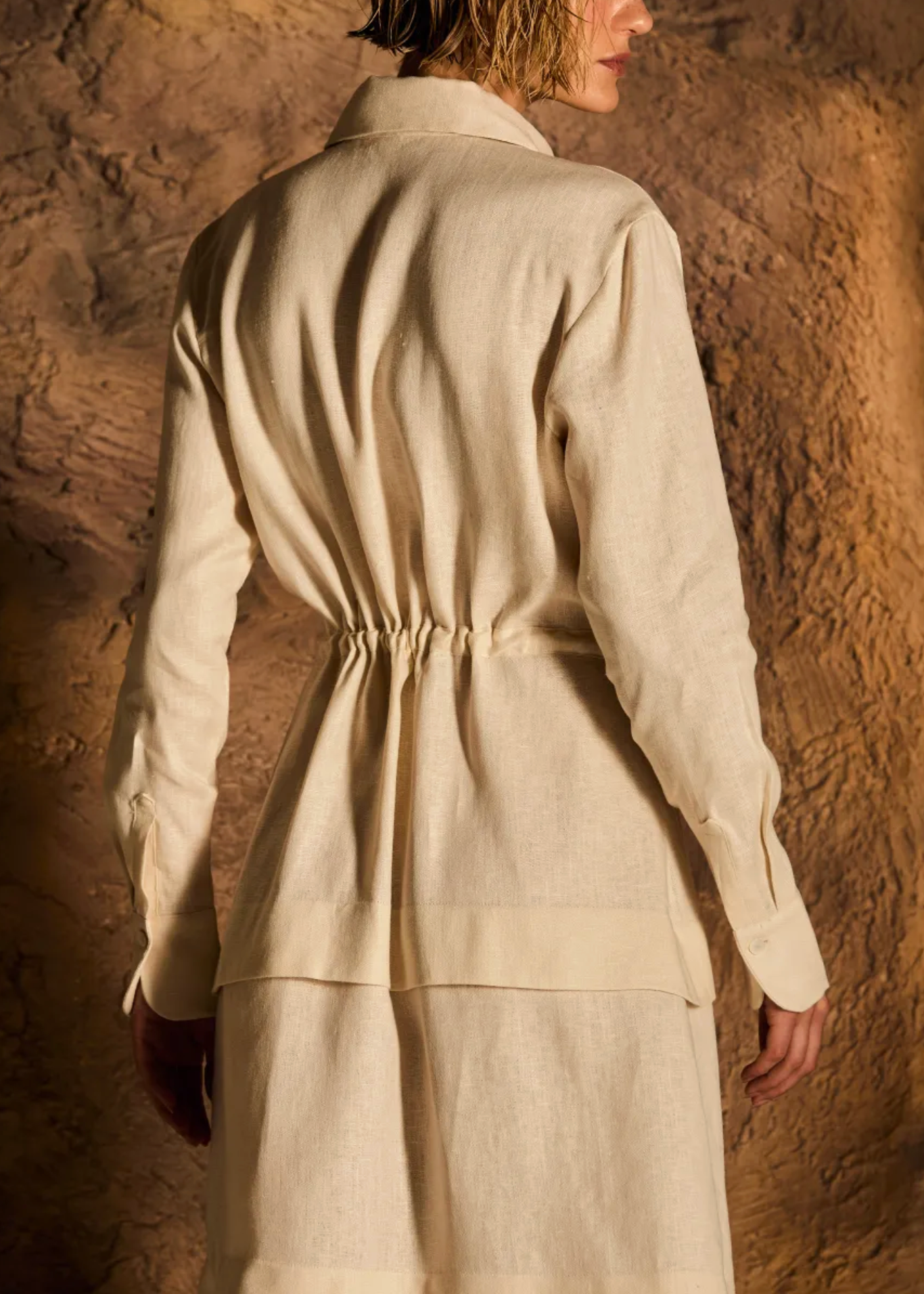 A model shown from the back wearing a cream-colored linen long-sleeve shirt and matching skirt set against a textured brown rock wall background.
