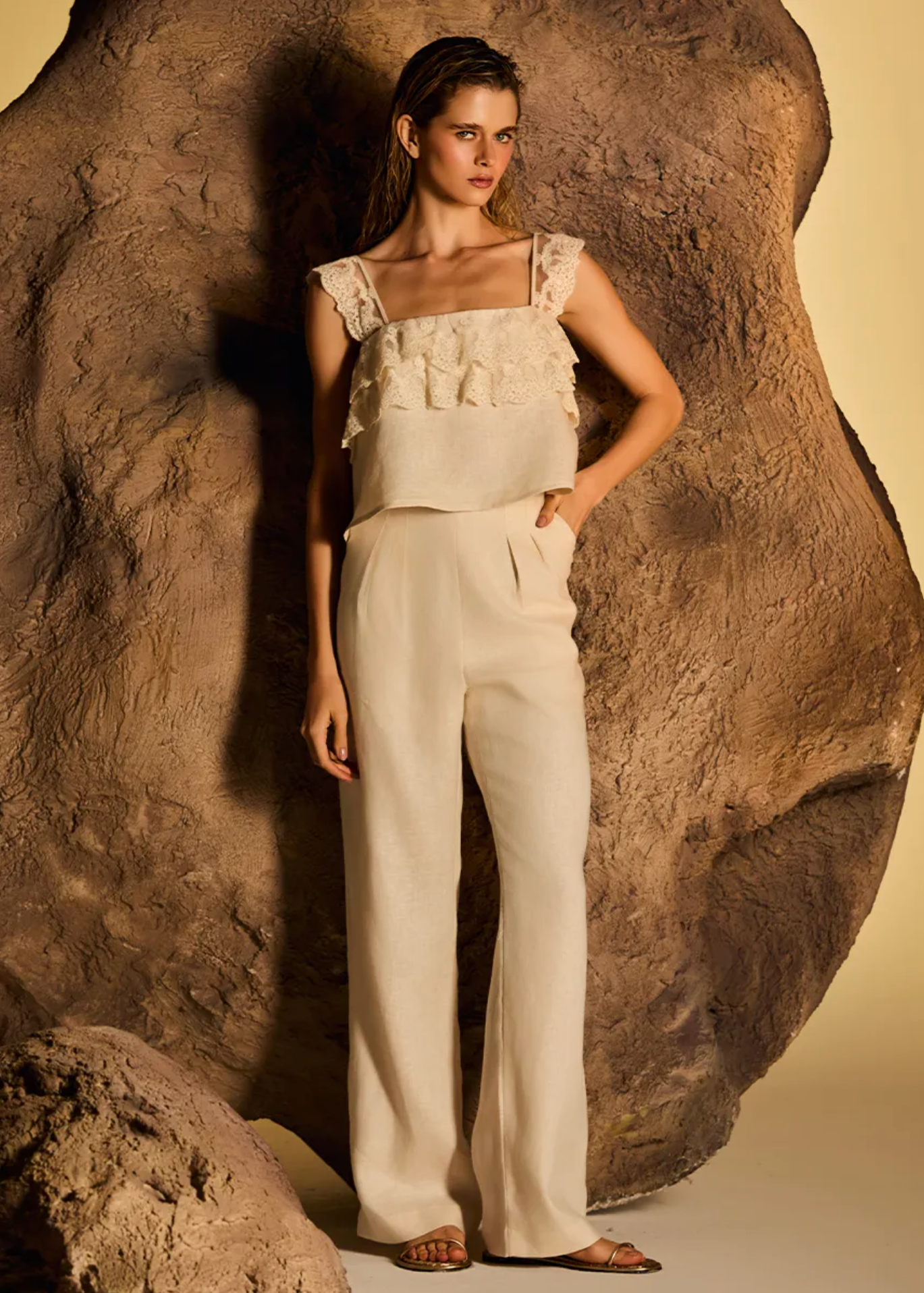 A model stands in front of a large textured rock prop wearing a cream-colored two-piece outfit consisting of a ruffled sleeveless top and matching wide-leg trousers.