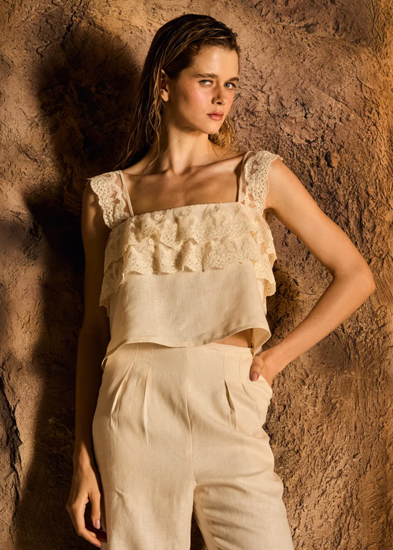 A model wearing a cream-colored two-piece set consisting of a ruffled lace-trimmed crop top and matching high-waisted trousers, posed against a textured brown rock wall.