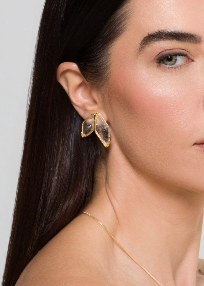 A close-up of a woman wearing a pair of gold geometric hoop earrings with clear crystal-like inlays, shown against a plain light gray background.