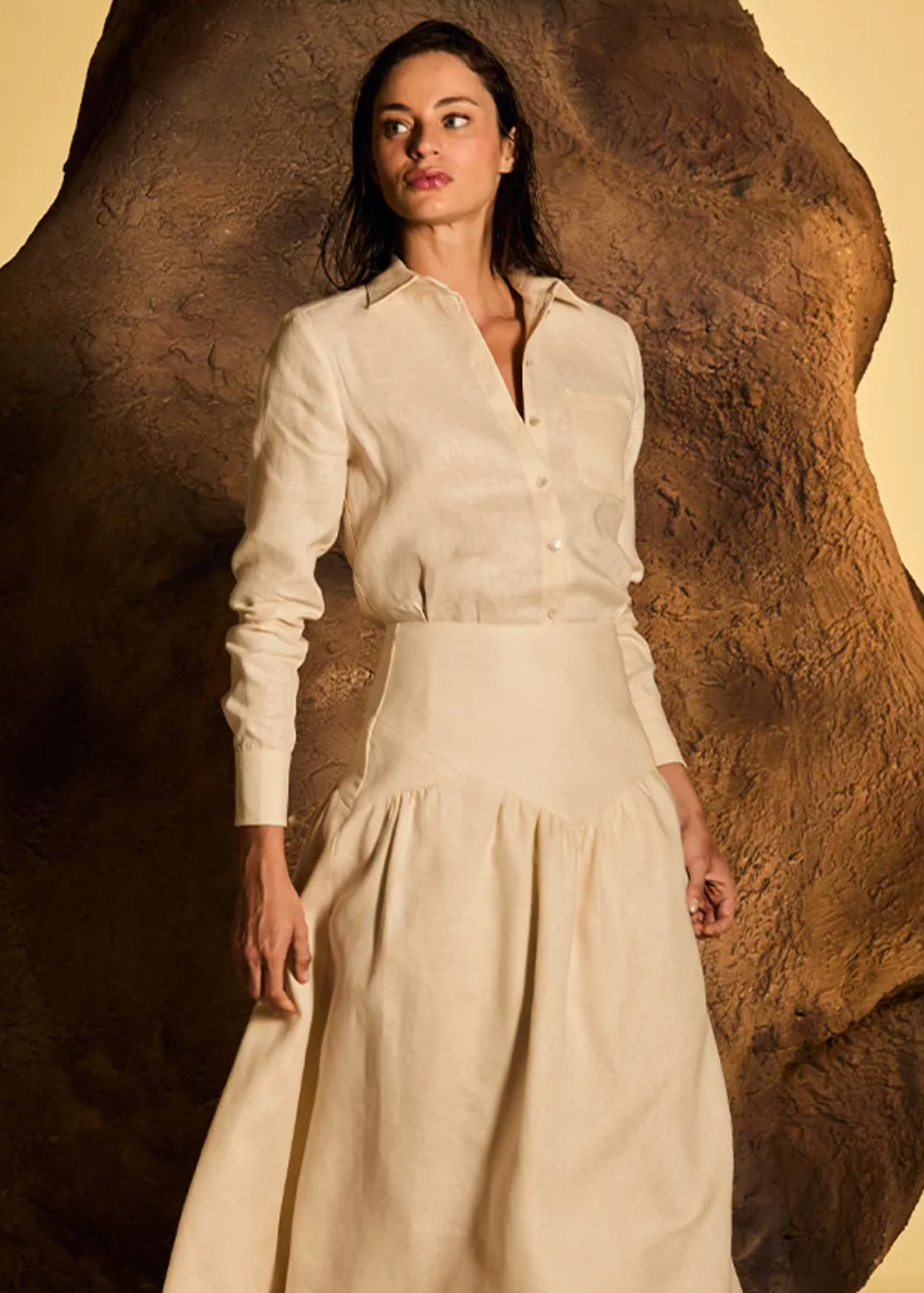 A woman models a cream-colored long-sleeve button-up shirt and matching high-waisted maxi skirt set, posed in front of a large textured brown rock prop against a warm beige background.