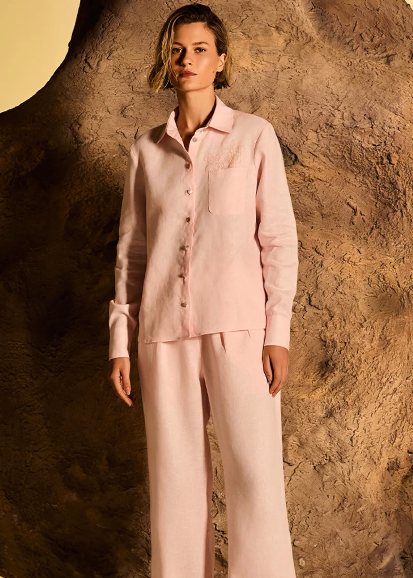 A woman modeling a light pink two-piece pajama set consisting of a long-sleeve button-up shirt and matching wide-leg pants, posed in front of a large textured brown rock formation.
