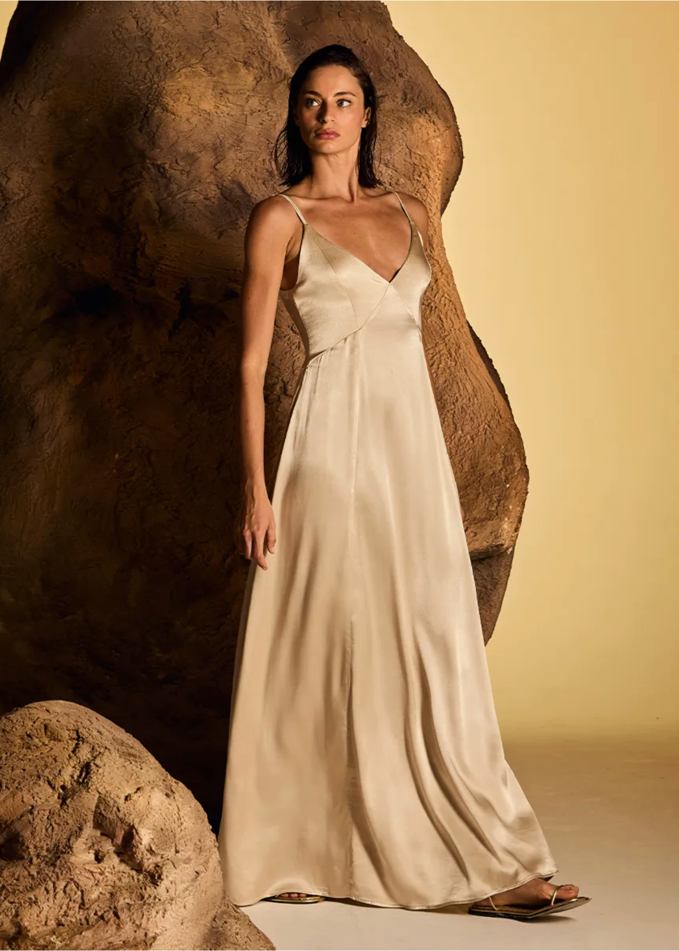 A woman wearing a floor-length satin slip dress in a warm cream or champagne tone, standing in front of a large sculptural rock against a beige studio backdrop.
