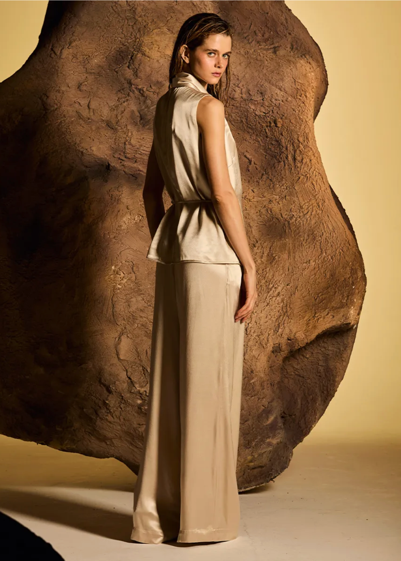 A model stands in profile wearing a sleeveless beige satin-like top and matching wide-leg pants, posed in front of a large sculptural rock against a warm beige studio background.