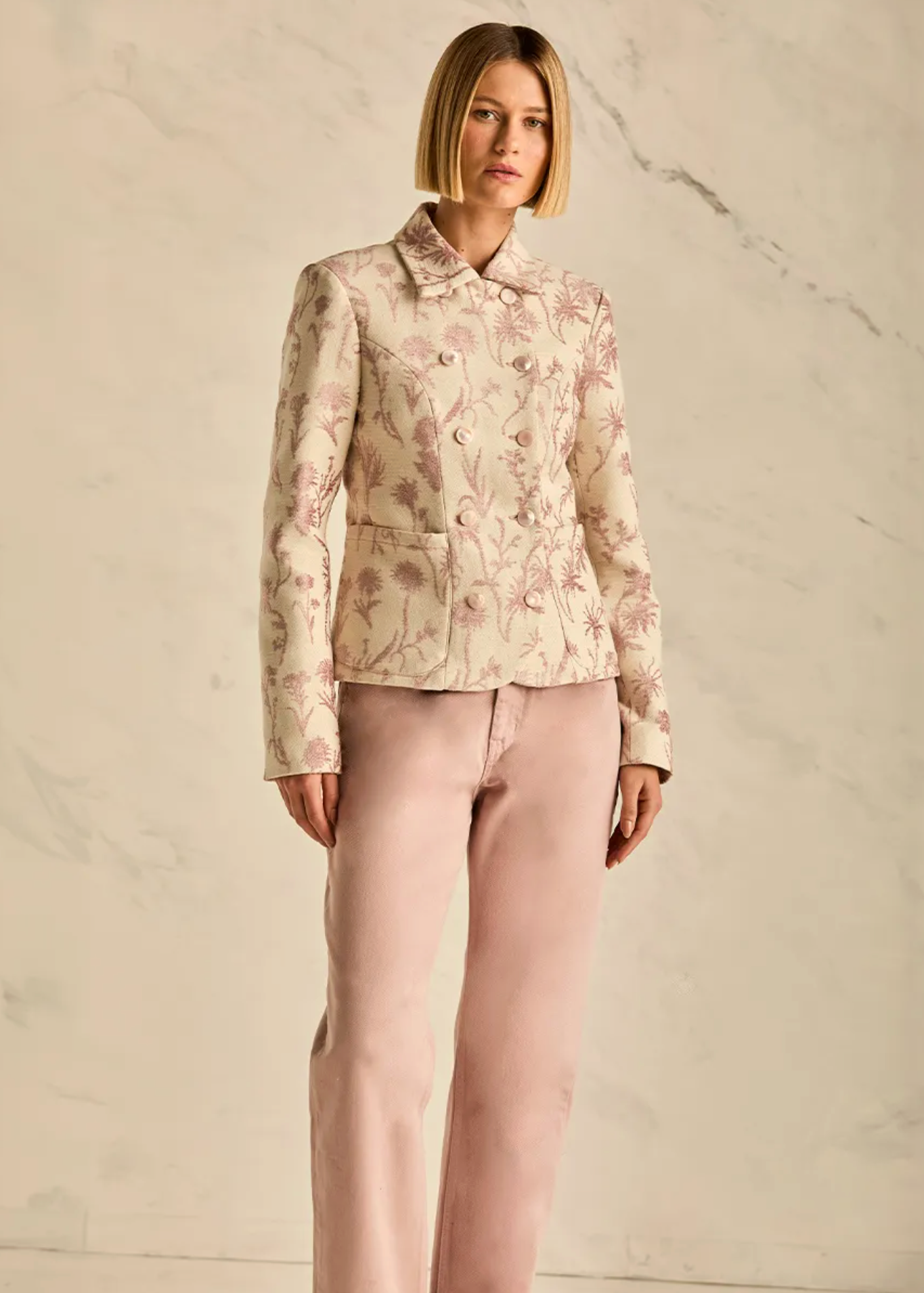 A model stands against a light beige marble-textured wall wearing a cropped, long-sleeve button-front jacket with a pink floral pattern on a cream base, paired with light pink trousers.