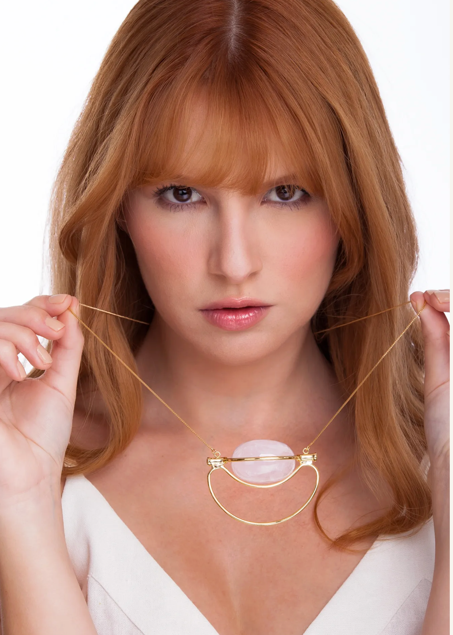 A woman with red hair wearing a white top holds a gold necklace with a crescent-shaped gold frame and a pale pink oval stone pendant against a white background.