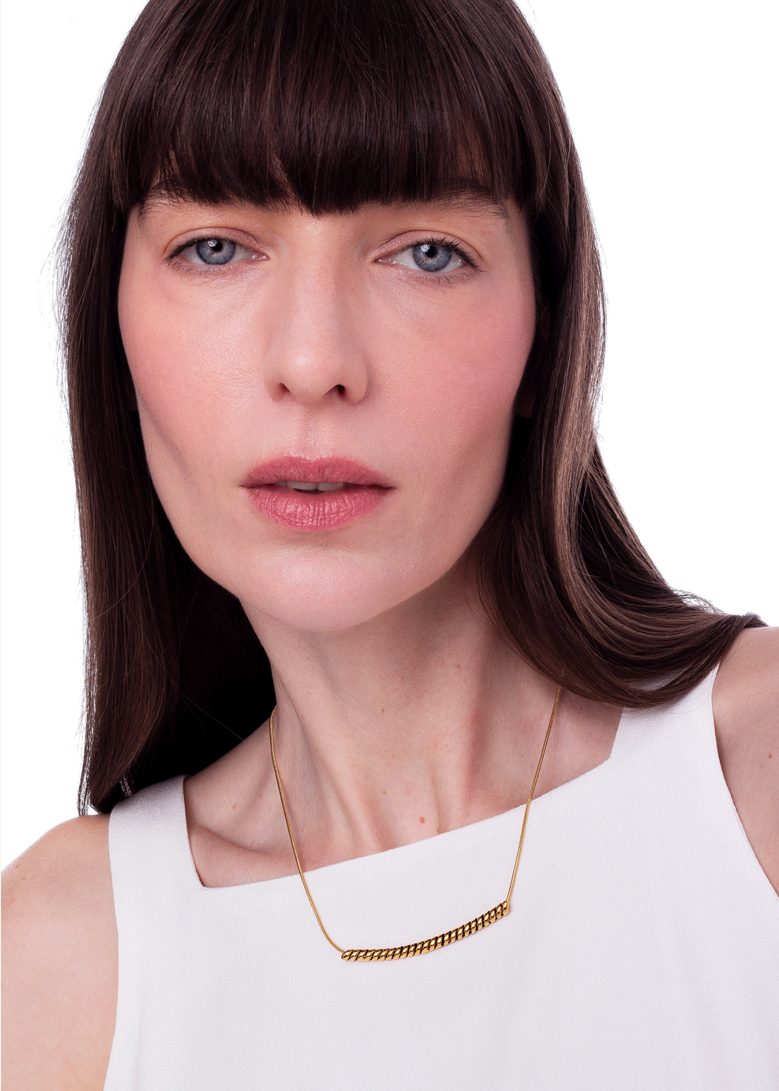 A close-up portrait of a woman with dark brown hair and bangs wearing a delicate gold chain necklace with a textured bar, paired with a white sleeveless top against a plain white background.