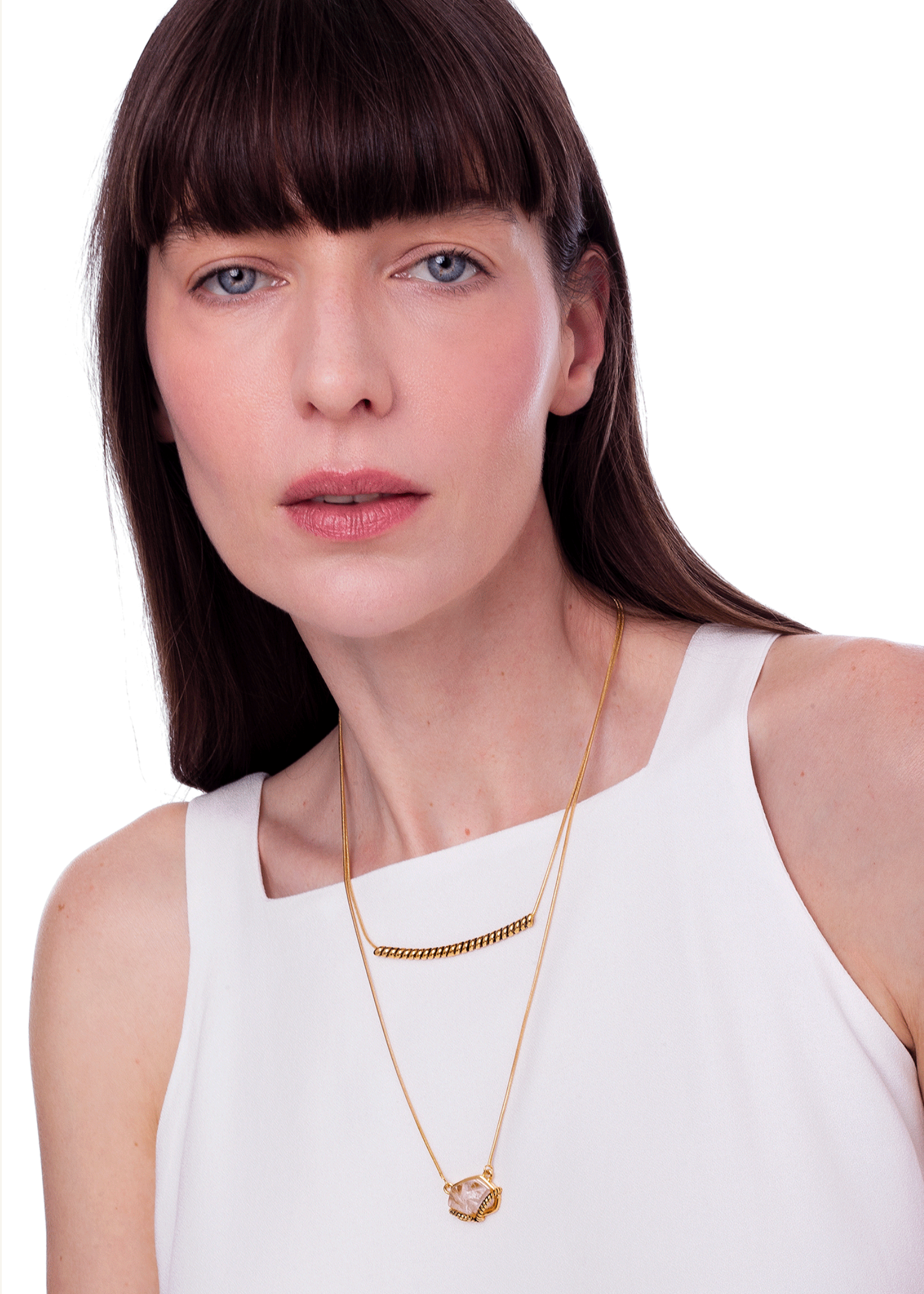 A woman with dark brown hair and bangs wearing a white sleeveless top, showcasing a layered gold necklace set against a plain white background.