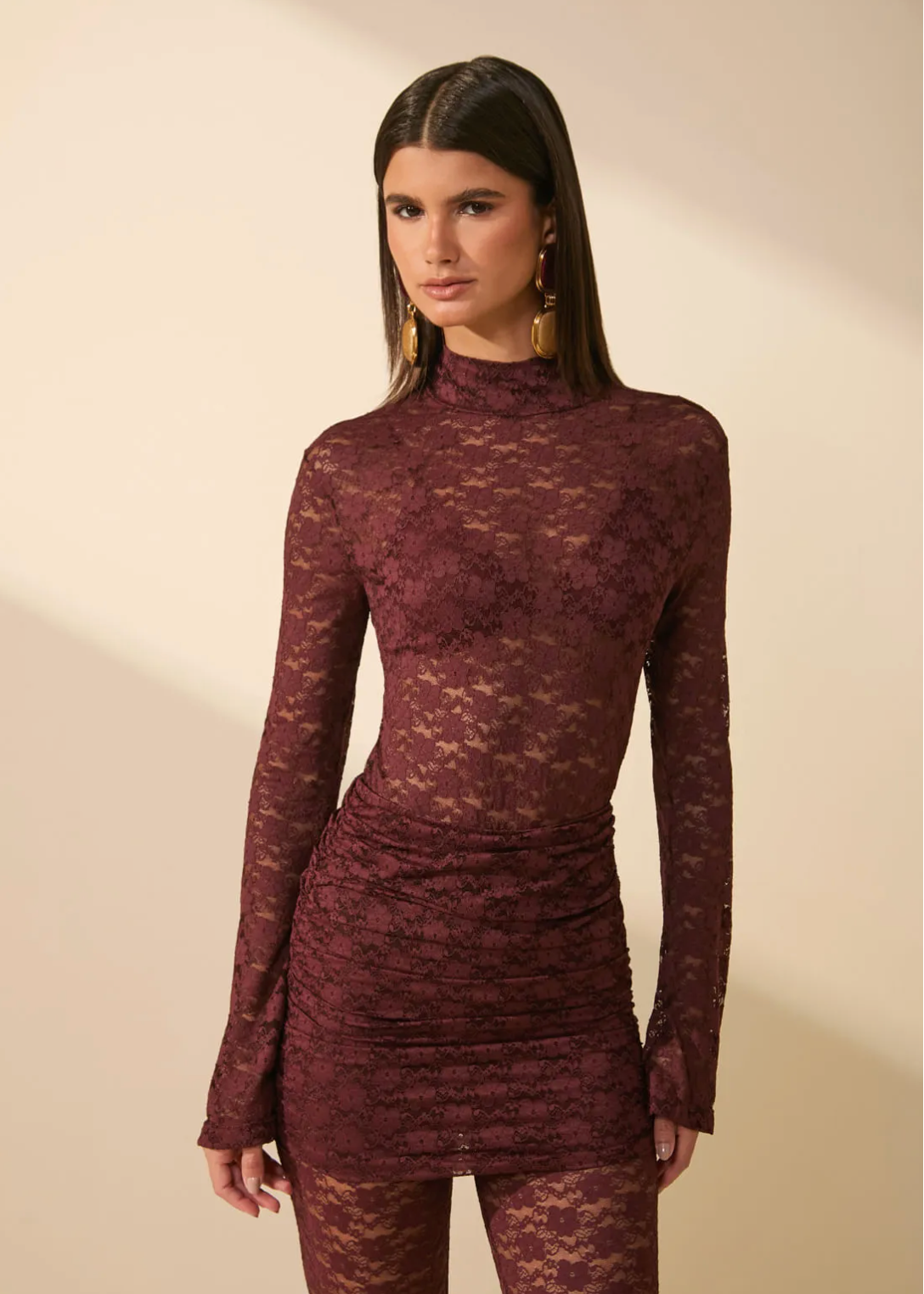 A woman modeling a long-sleeve, high-neck, sheer lace mini dress in a deep burgundy shade, paired with matching lace tights, set against a warm beige background with soft diagonal shadows.