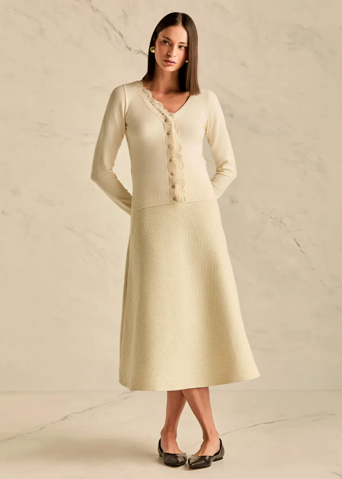 A woman modeling a cream-colored knit midi dress with a textured skirt and a lace-trimmed button-front bodice, standing against a light beige marble wall and floor.