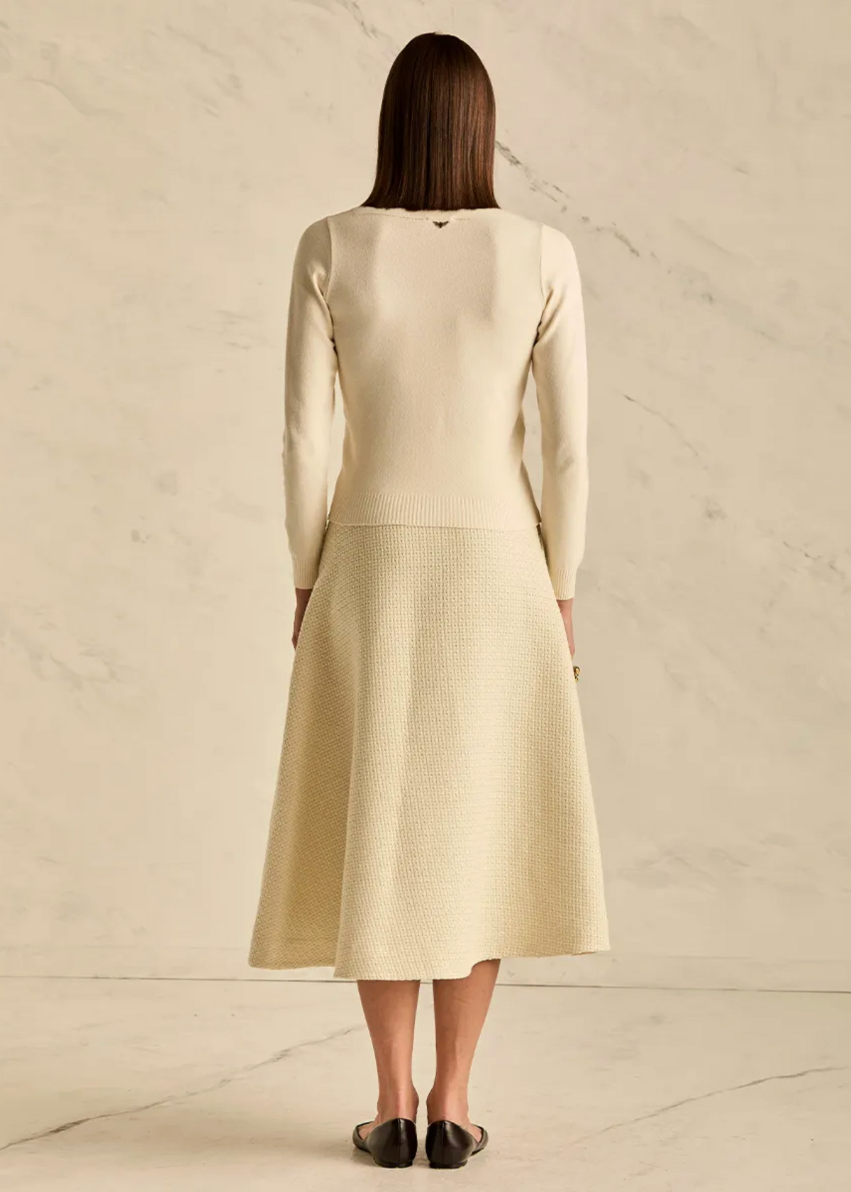 A woman viewed from behind wearing a cream-colored knit long-sleeve top and matching textured midi skirt, standing against a light beige marble wall and floor.