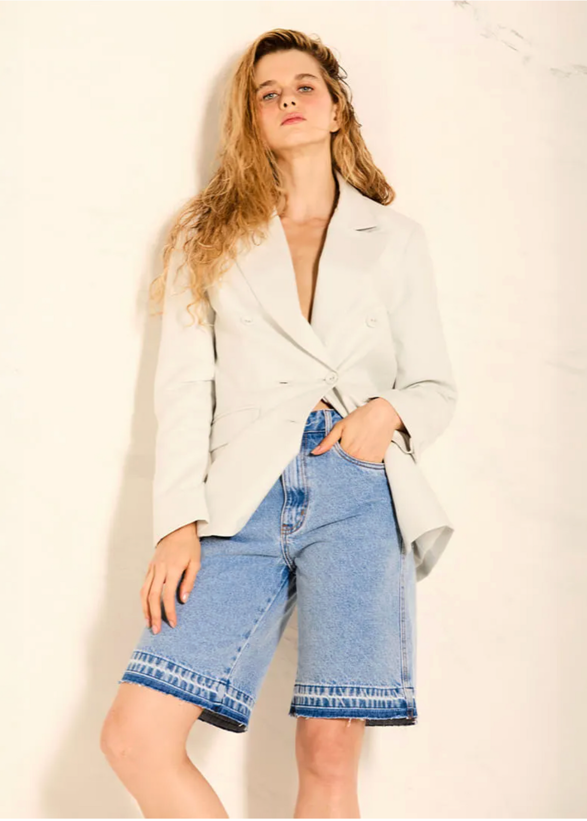 A woman posing in a light gray double-breasted blazer and light-wash denim shorts against a textured off-white wall.