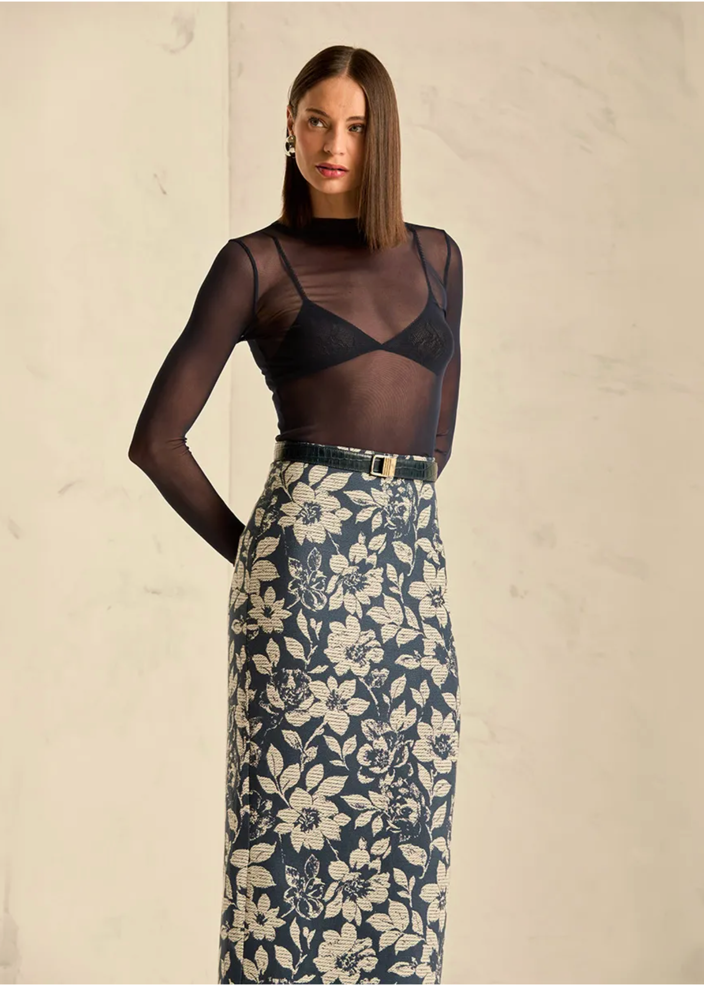 A model stands against a light beige textured wall wearing a sheer black long-sleeve mesh top over a black bralette, paired with a high-waisted navy floral skirt featuring a cream floral pattern and a black belt with a gold buckle.