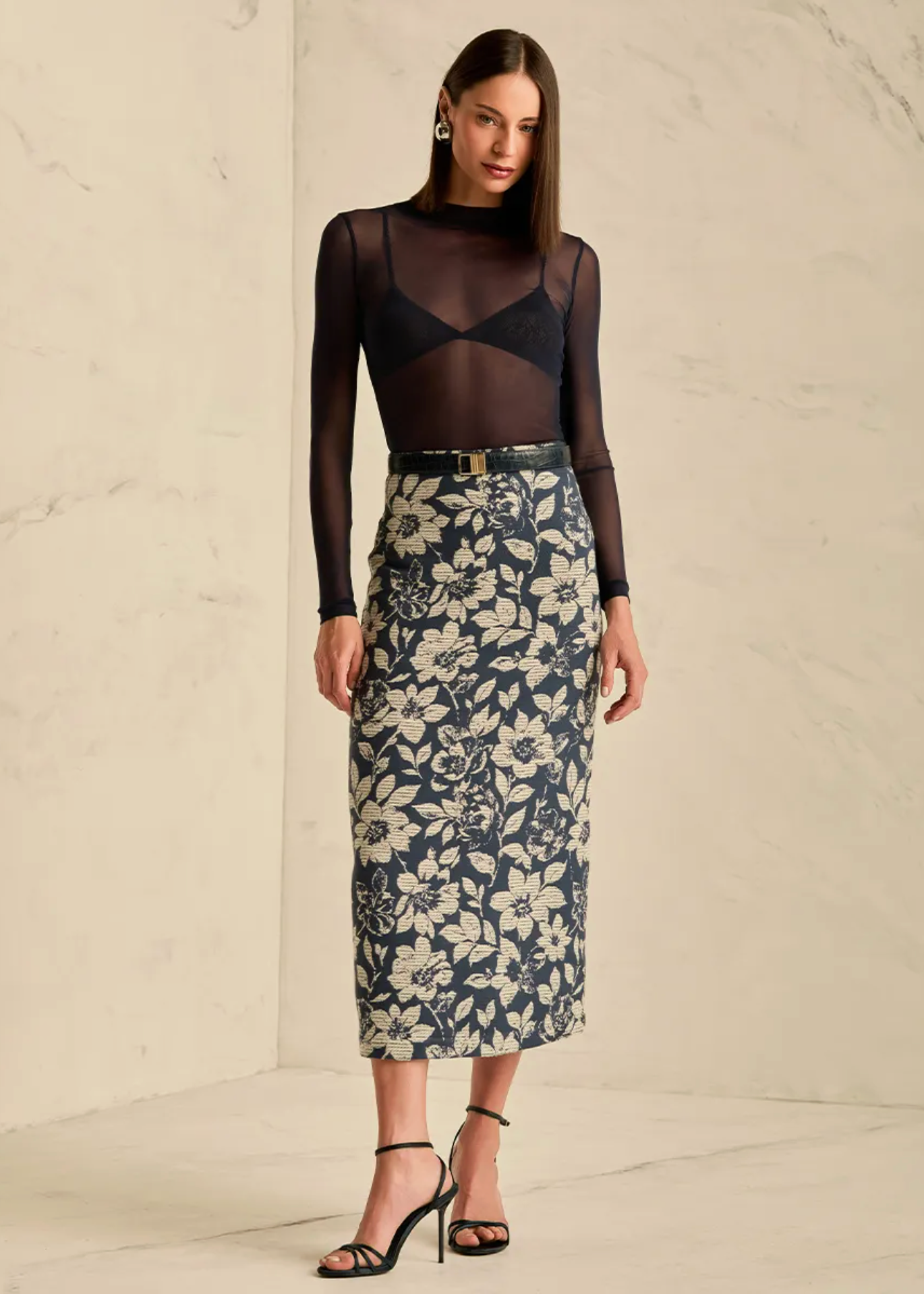 A model stands in a studio setting wearing a sheer black long-sleeve mock-neck top paired with a navy blue midi skirt featuring a cream floral pattern, accessorized with a thin belt and black strappy heels.