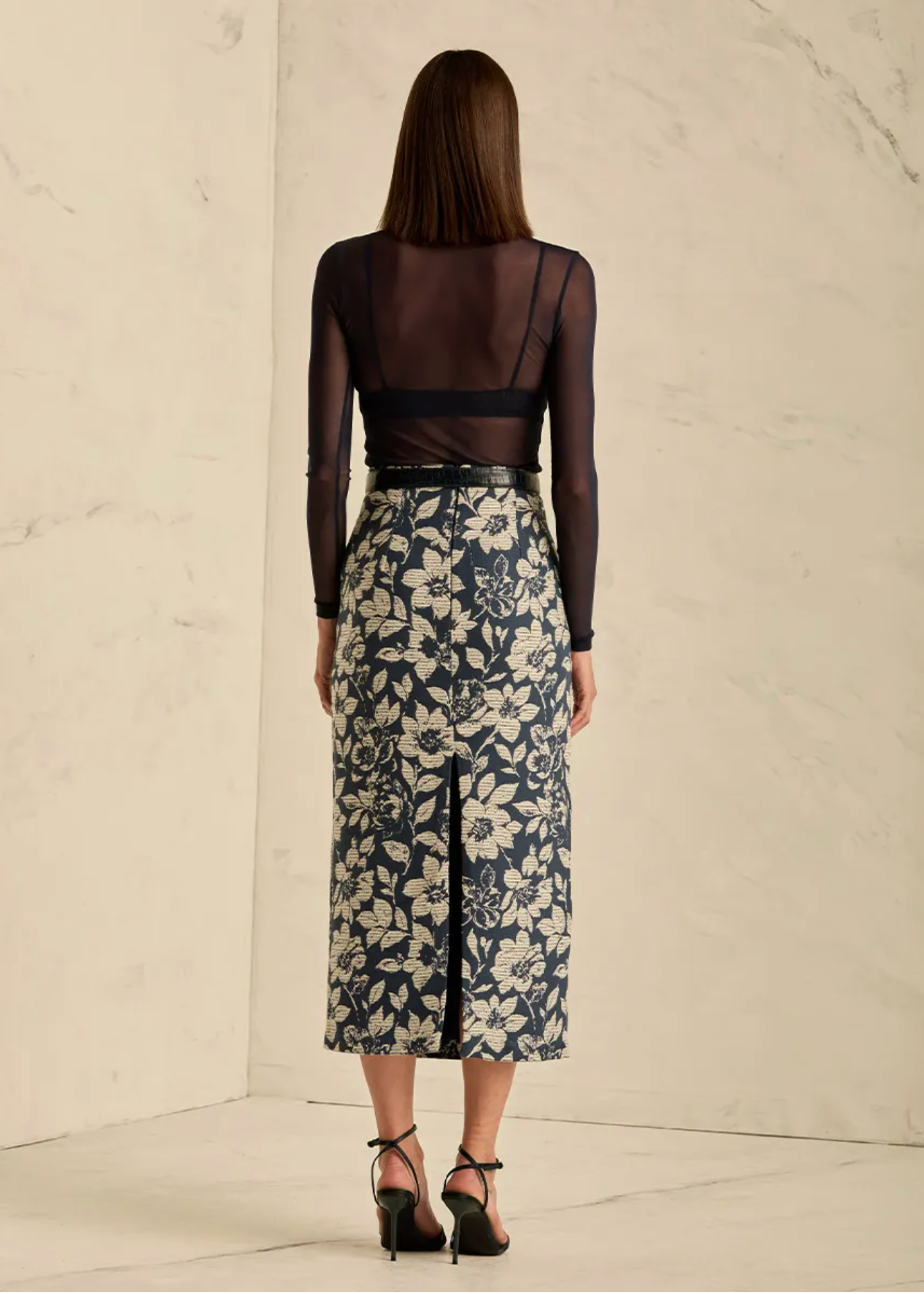 A woman stands with her back to the camera wearing a sheer black long-sleeve top over a navy floral midi skirt with a front slit, paired with black heels, against a light marble wall.