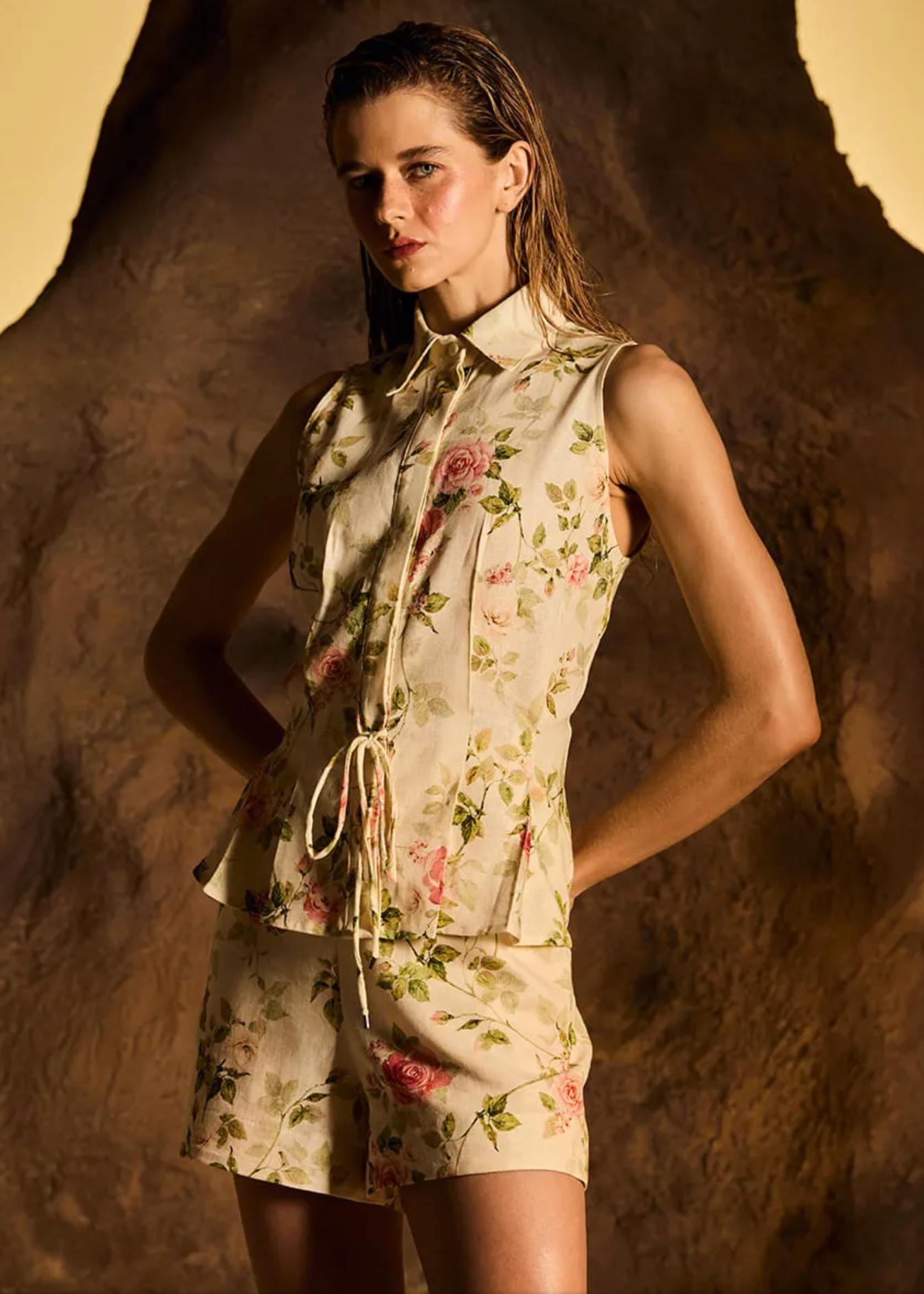 A woman models a sleeveless collared top and matching shorts set in a cream floral print with pink roses and green leaves, posed against a textured brown rock backdrop with dramatic warm lighting.
