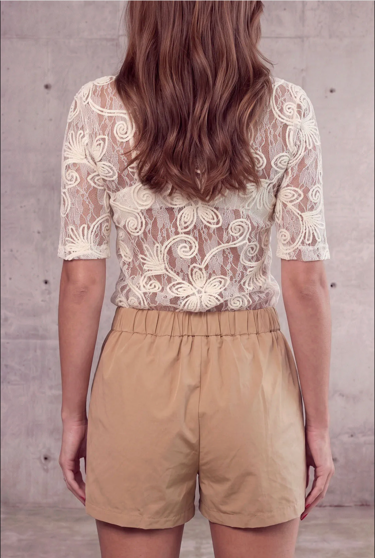 A woman shown from the back wearing a cream lace short-sleeve top and tan shorts, standing against a textured concrete wall.