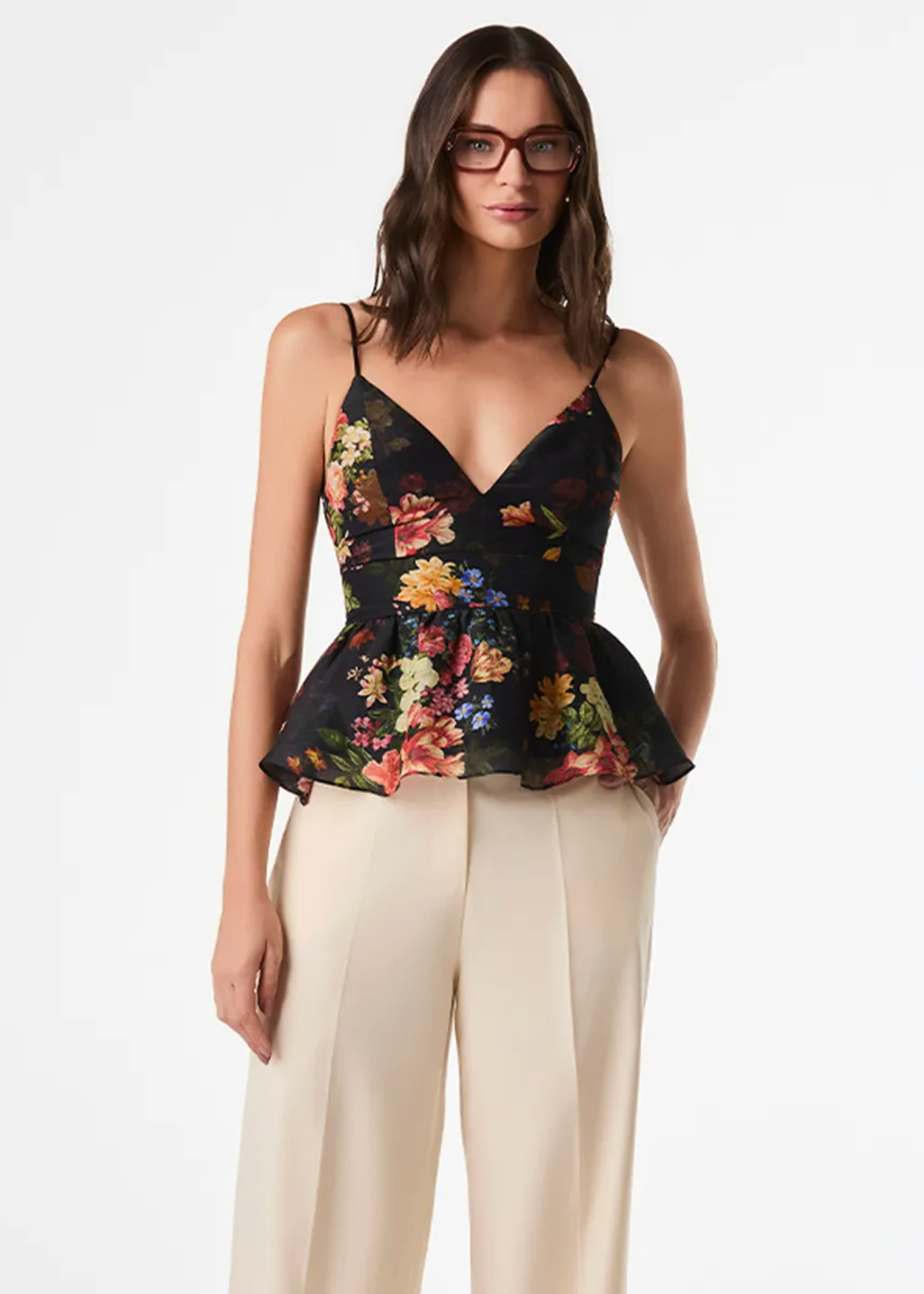 A woman modeling a black floral camisole top with thin straps and a peplum hem, paired with cream wide-leg pants, against a plain white studio background.