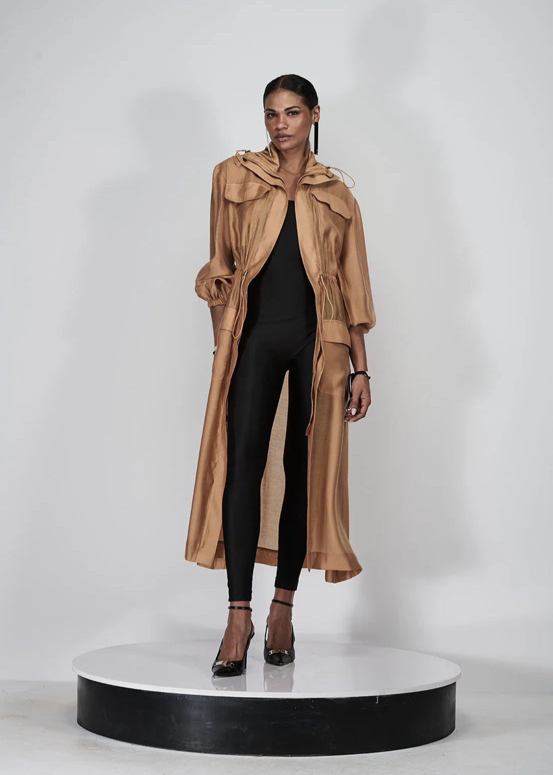 Model wearing a long beige coat over a black outfit on a white background