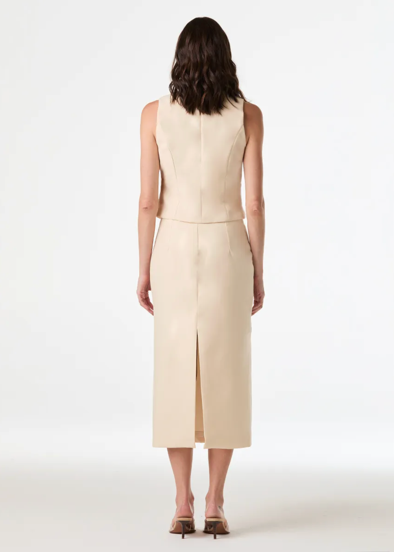 A woman stands with her back to the camera wearing a sleeveless, structured midi dress in a light cream or beige color. The dress features a straight silhouette, a high neckline, and a front slit at the hem.