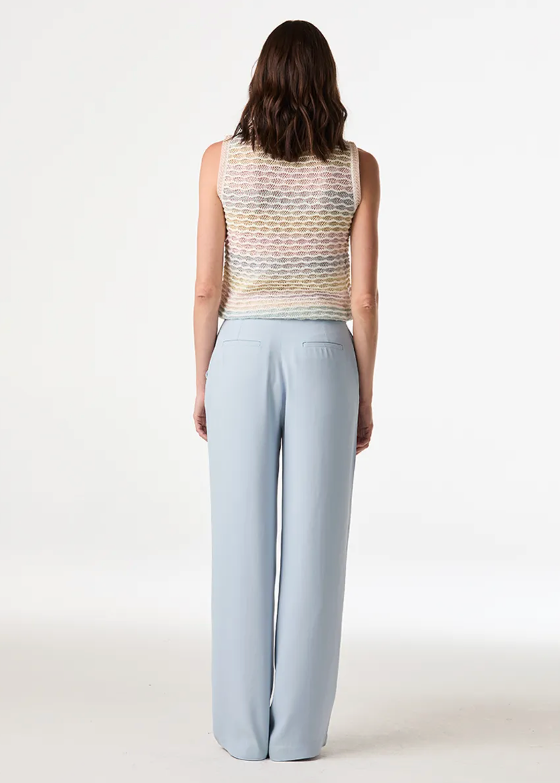 A woman shown from the back wearing a sleeveless knit top with a multicolor wavy pattern and light blue wide-leg trousers, photographed against a clean white studio background.