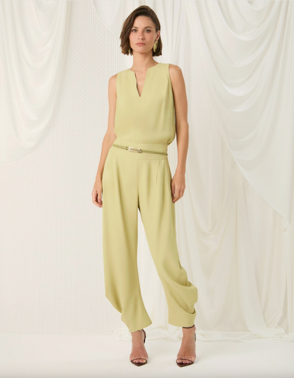 A woman models a sleeveless light yellow-green jumpsuit with a V-neckline, wide legs, and a matching belt with a silver buckle, set against a draped white fabric backdrop.