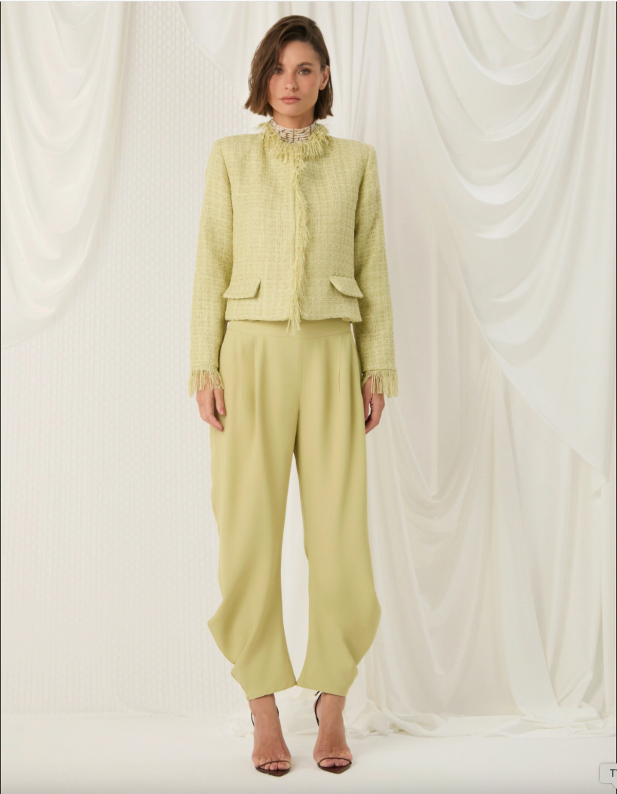 A model stands against a draped white fabric backdrop wearing a pale yellow-green tweed jacket with fringe trim and matching wide-leg trousers.