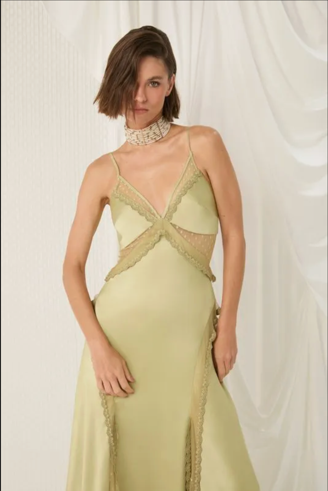 A woman models a pale yellow-green satin gown with beige lace trim and a crossover criss-cross bodice detail, styled with a beaded choker against a draped white fabric background.