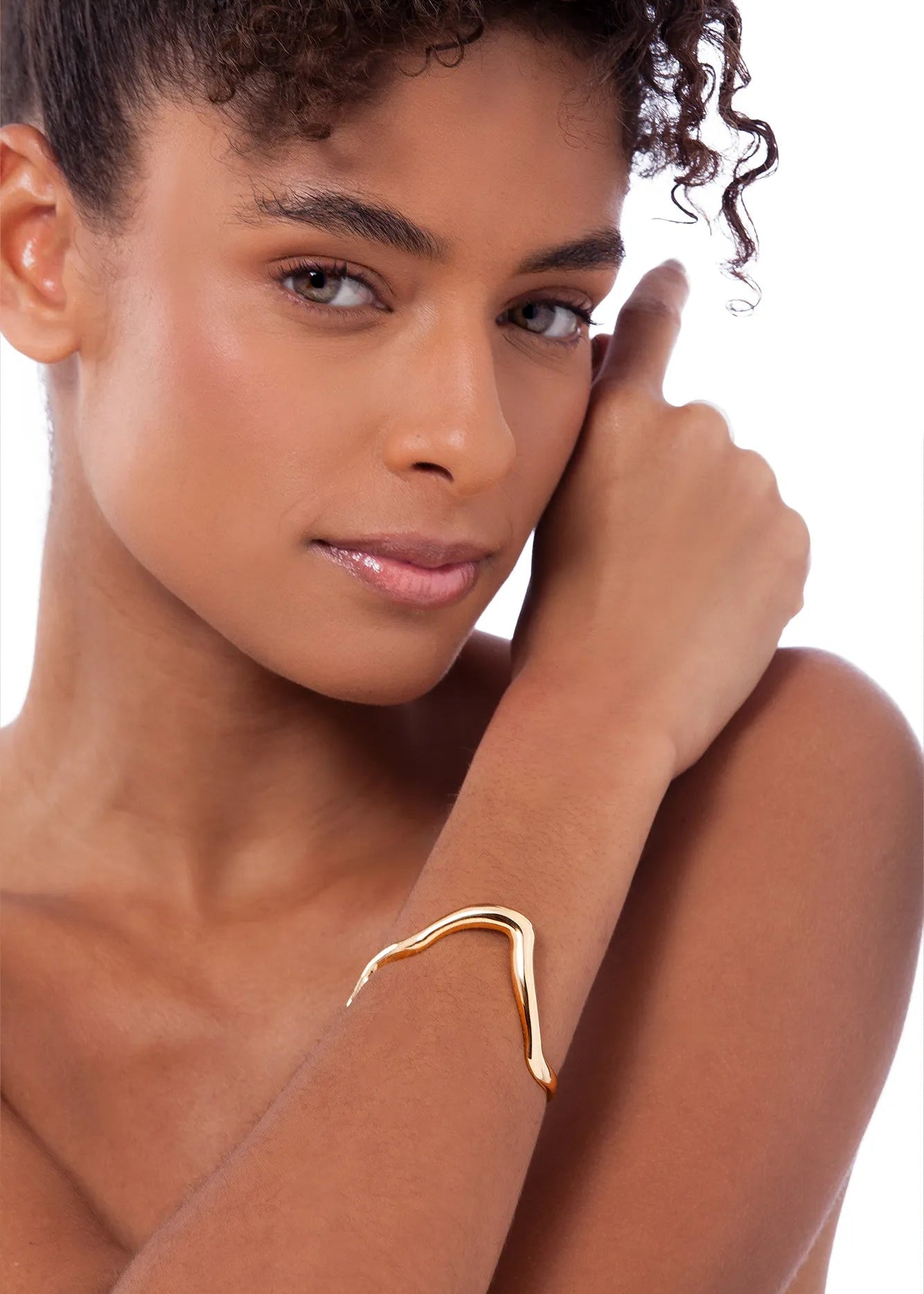 A close-up of a model wearing a thin, wavy gold cuff bracelet on her wrist, set against a plain white background.