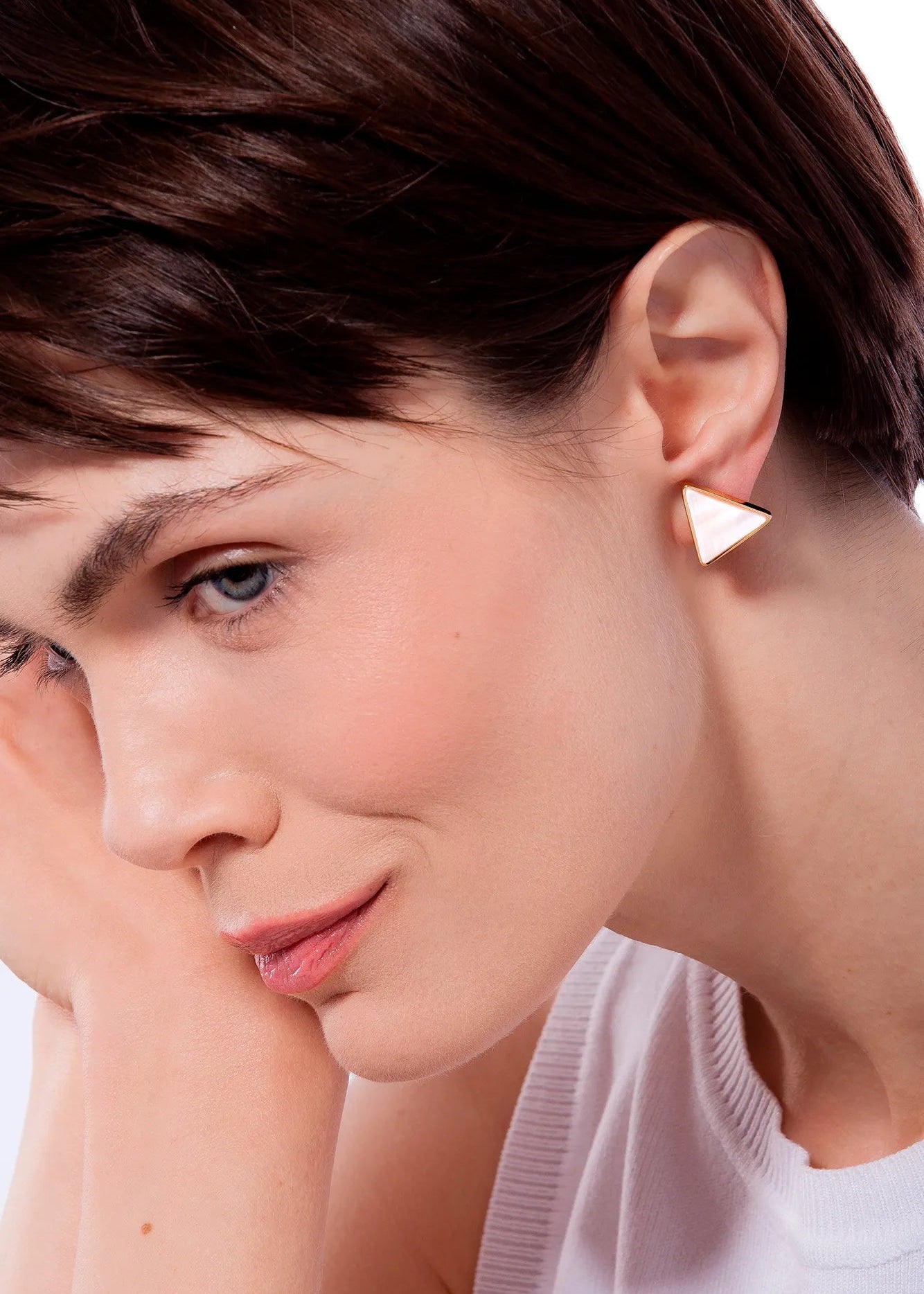 A close-up of a woman with short brown hair wearing a small white triangular stud earring, resting her chin on her hand against a white background.