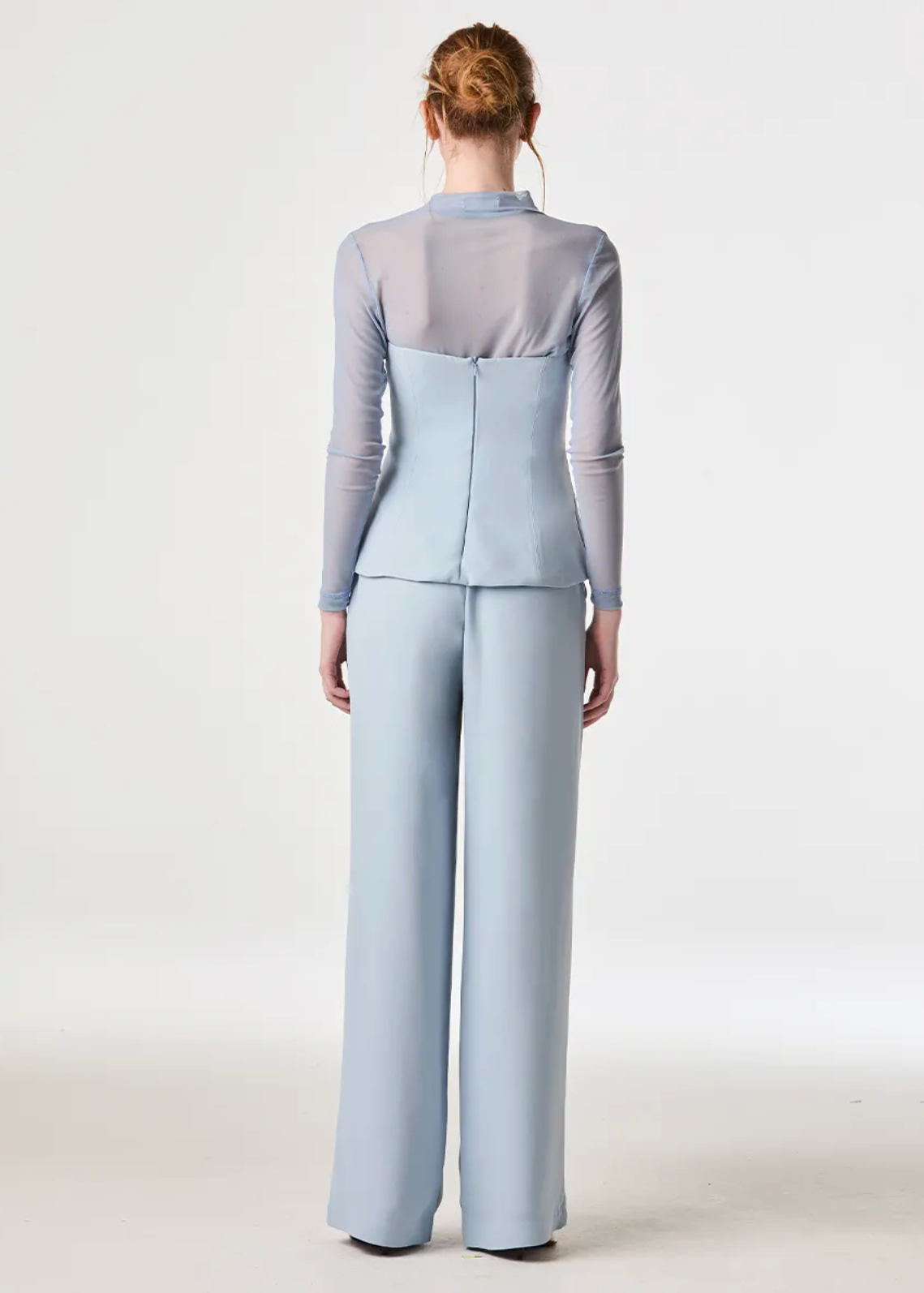 A model viewed from the back wearing a light blue two-piece outfit consisting of a long-sleeve top with a sheer yoke and matching wide-leg trousers, against a plain white studio background.