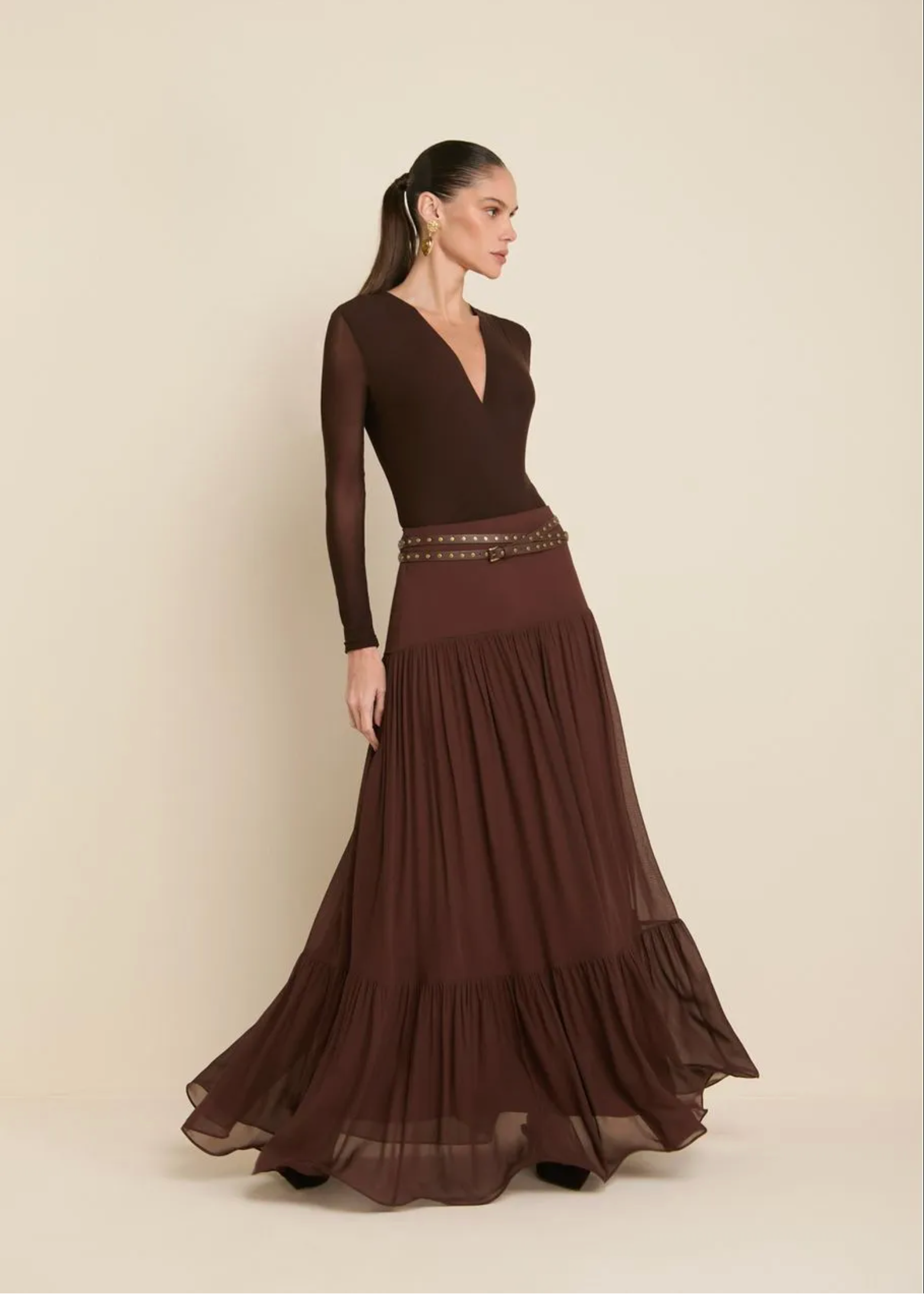 A woman modeling a long-sleeve maxi dress in a deep chocolate brown color, featuring a structured bodice, a thin studded waist belt, and a flowing tiered chiffon skirt, set against a plain beige studio background.