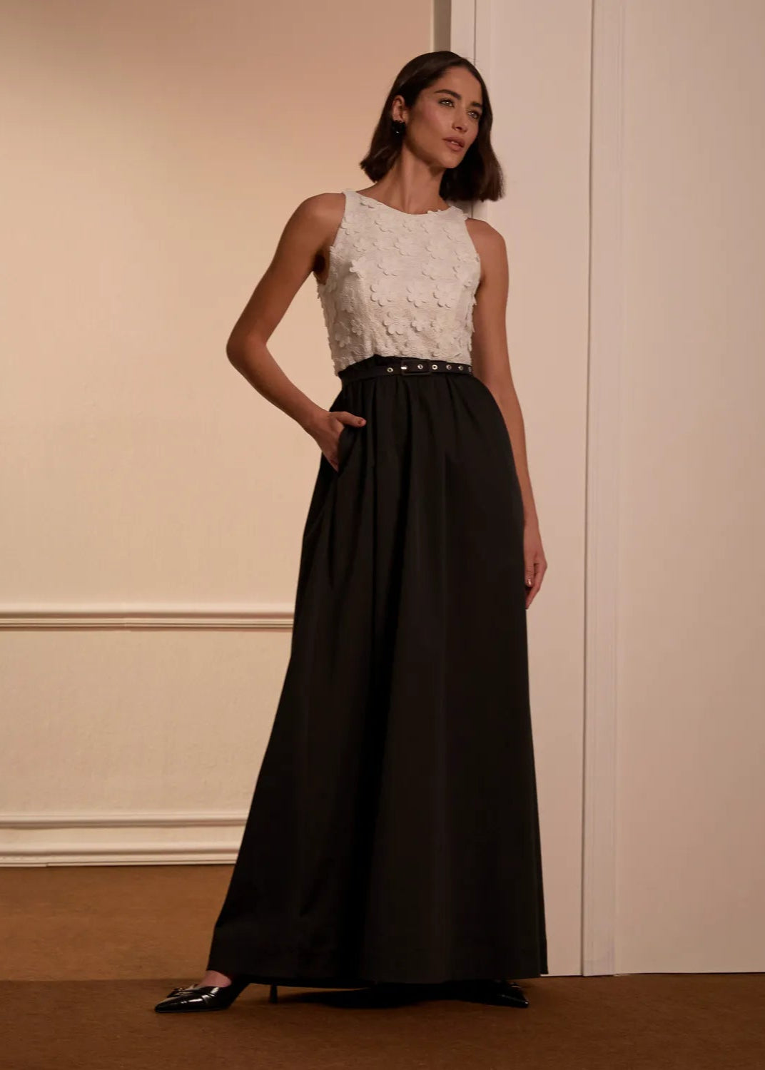 Woman wearing a white lace top and black skirt against a beige wall.