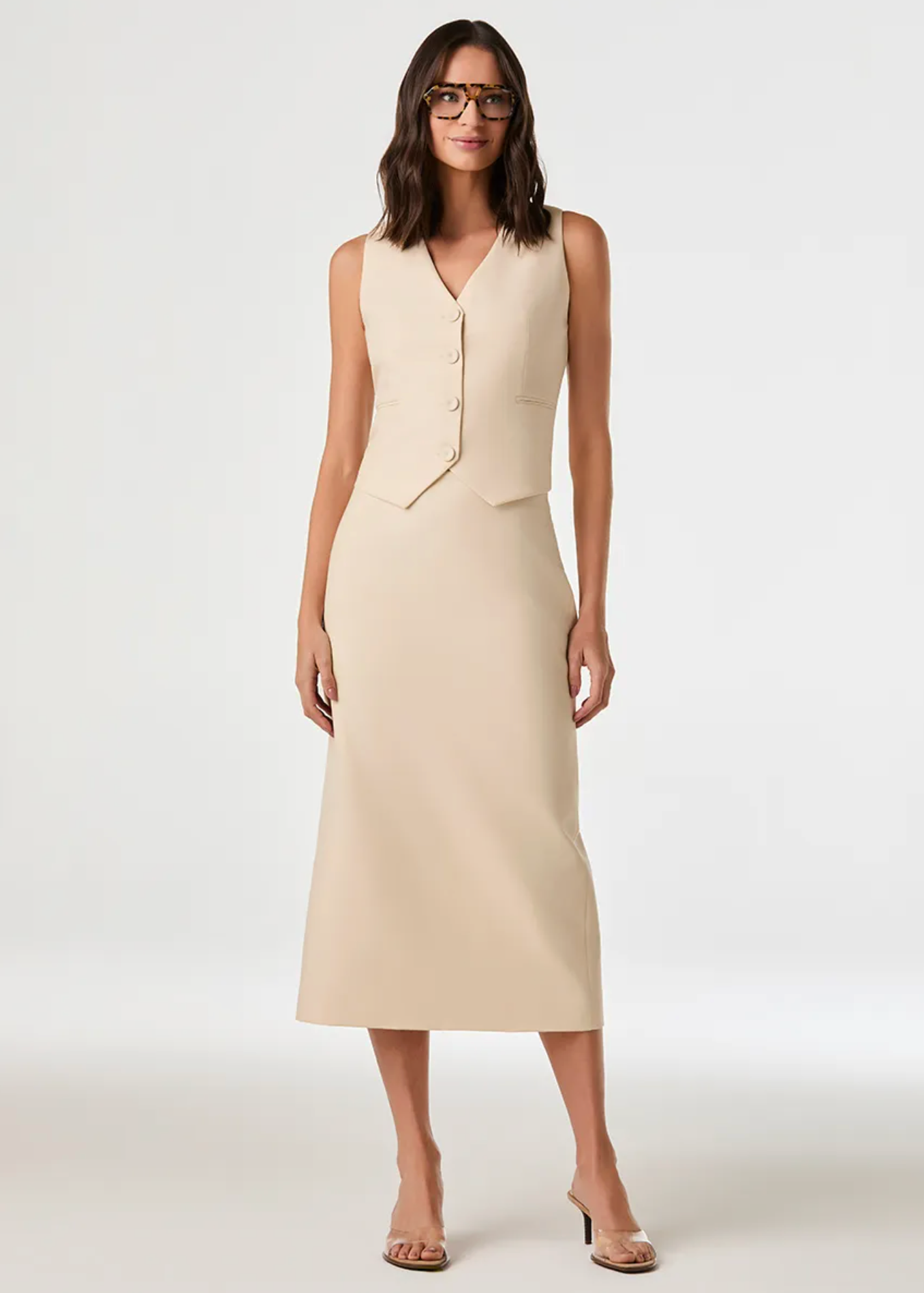 A woman modeling a sleeveless beige midi dress with a structured vest-like bodice and front buttons, photographed against a clean light gray studio background.
