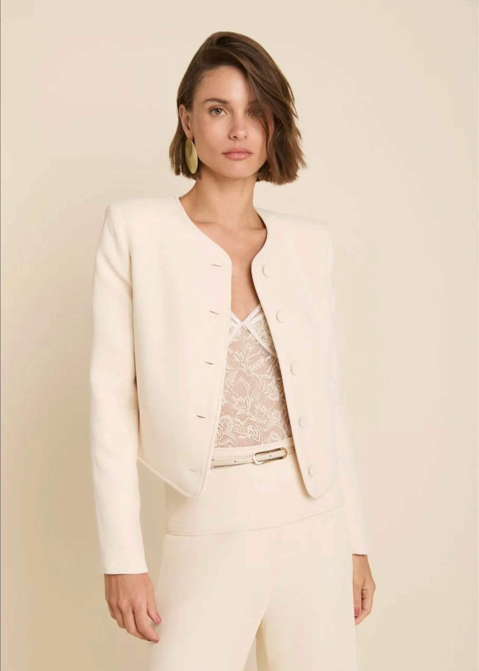 A woman modeling a cream-colored cropped blazer with decorative buttons, paired with matching trousers and a lace top, set against a solid light beige background.