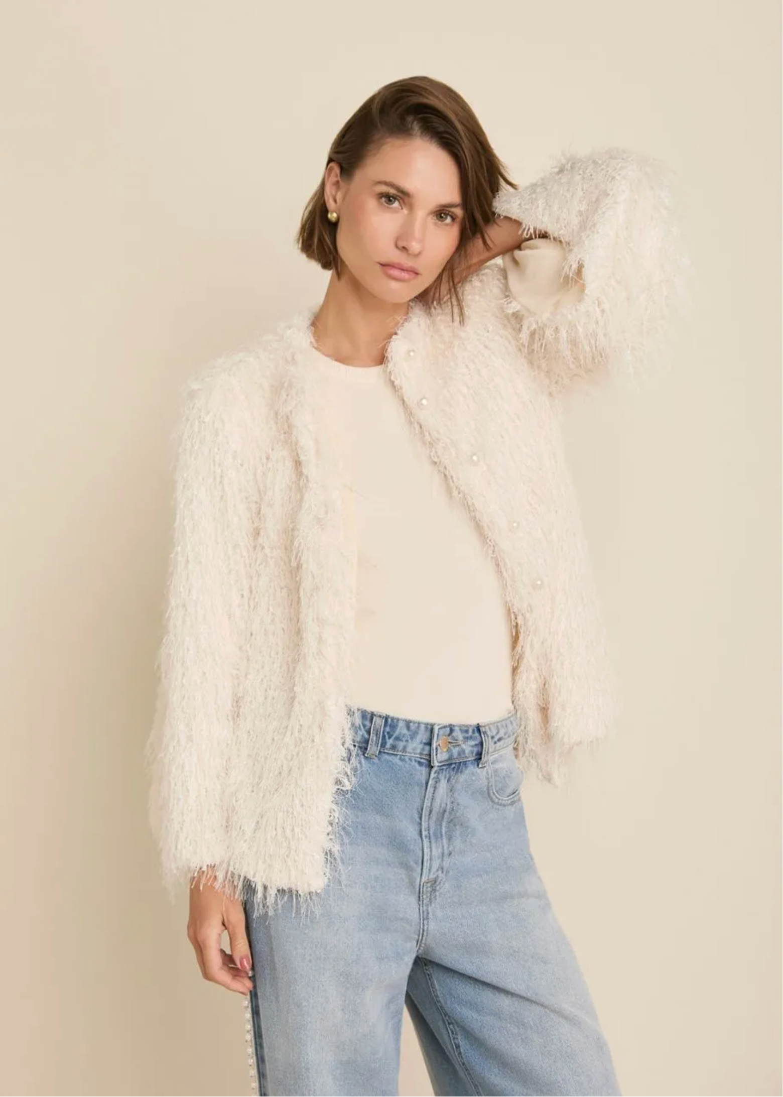 A woman modeling a fluffy cream-colored open-front jacket over a cream top and light blue jeans, set against a solid light beige studio background.